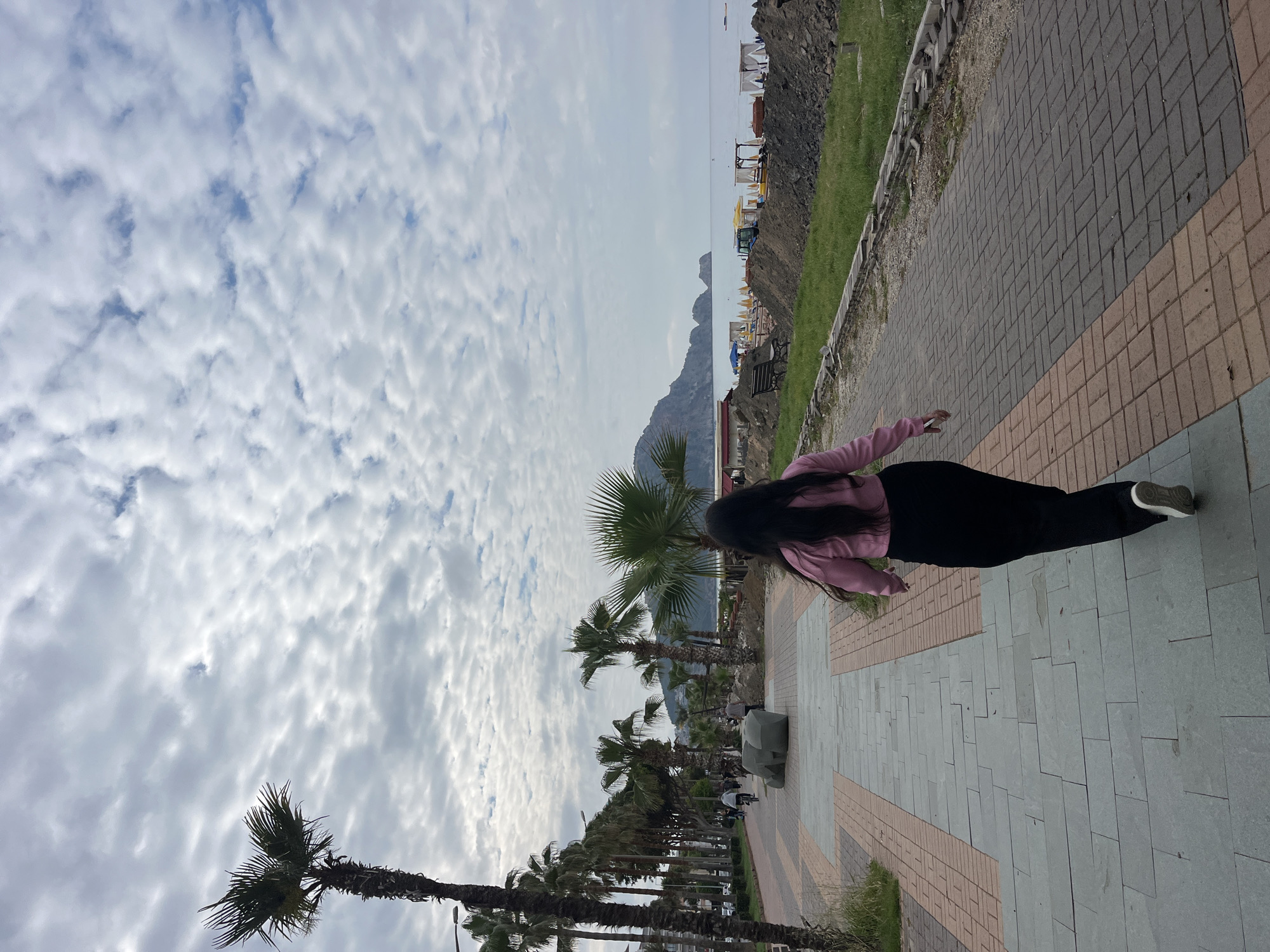 The photograph, taken outdoors, captures a scene along a pedestrian walkway in Alanya, Turkey. A person is positioned on the walkway, wearing a pink long-sleeved shirt and black pants, with dark hair. The walkway is constructed of gray and reddish-brown rectangular paving stones. To the left, palm trees line the walkway, framing a view of a mountain range and a sky filled with layered clouds. To the right, there is a grassy area, followed by a stone embankment, and a beach area with yellow canopies. The image appears to be shot during daylight hours, with overcast skies.