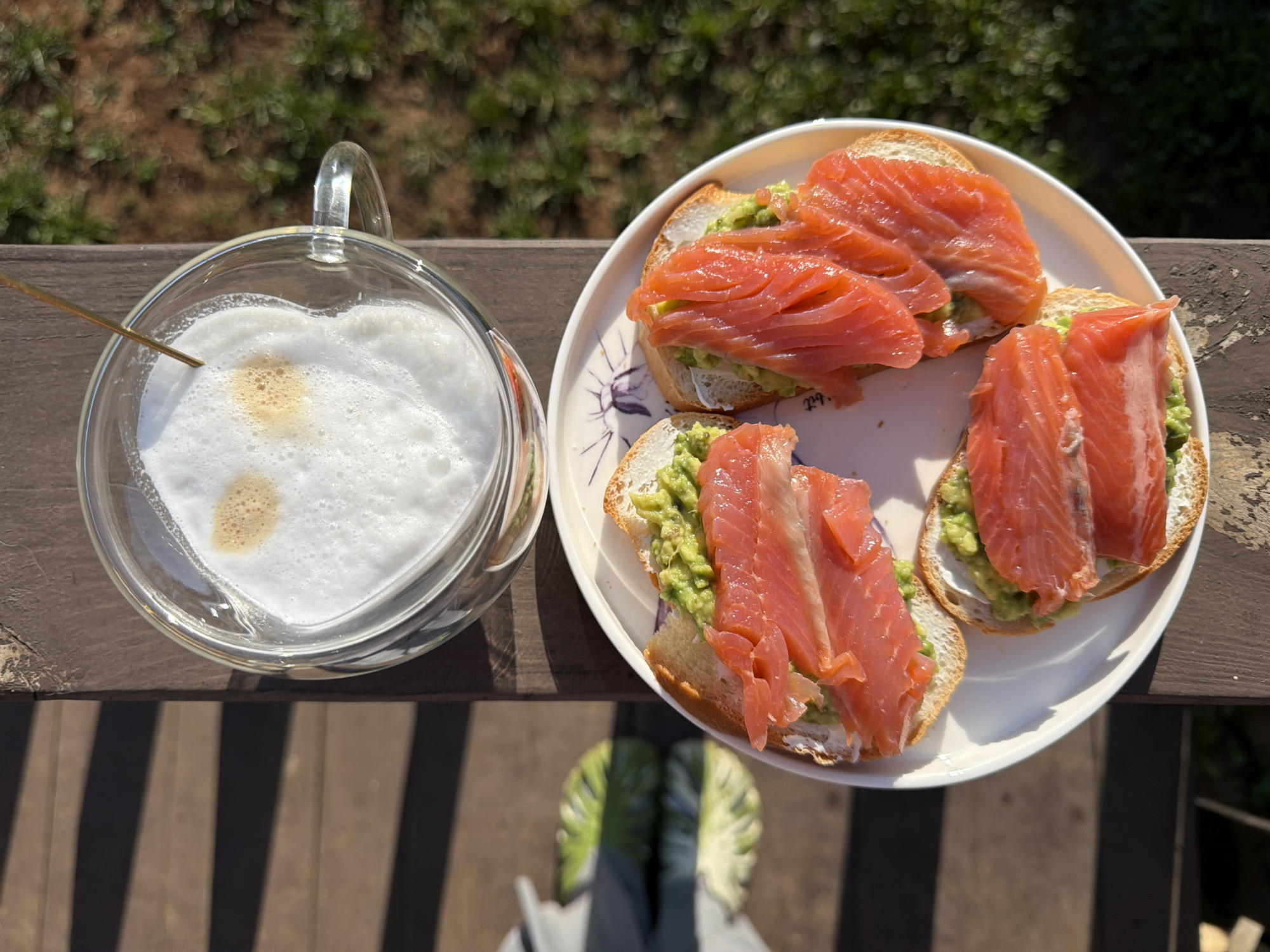 The image shows a close-up, overhead view of a meal and beverage placed on a wooden surface, possibly a deck. A heart-shaped glass mug filled with a white, foamy beverage (likely a latte or cappuccino) with two small spots of brown liquid on the surface is positioned to the left. To the right, a white plate holds three slices of bread topped with green avocado and slices of salmon. The background shows blurred greenery, suggesting an outdoor setting. In the lower portion of the image, two feet wearing sneakers with green and white detailing can be seen. The scene appears to be captured in daylight, with strong shadows indicating a sunny day. The location is unspecified, but the prompt suggests Chaikovsky, Russia. No specific information indicates that location is confirmed.