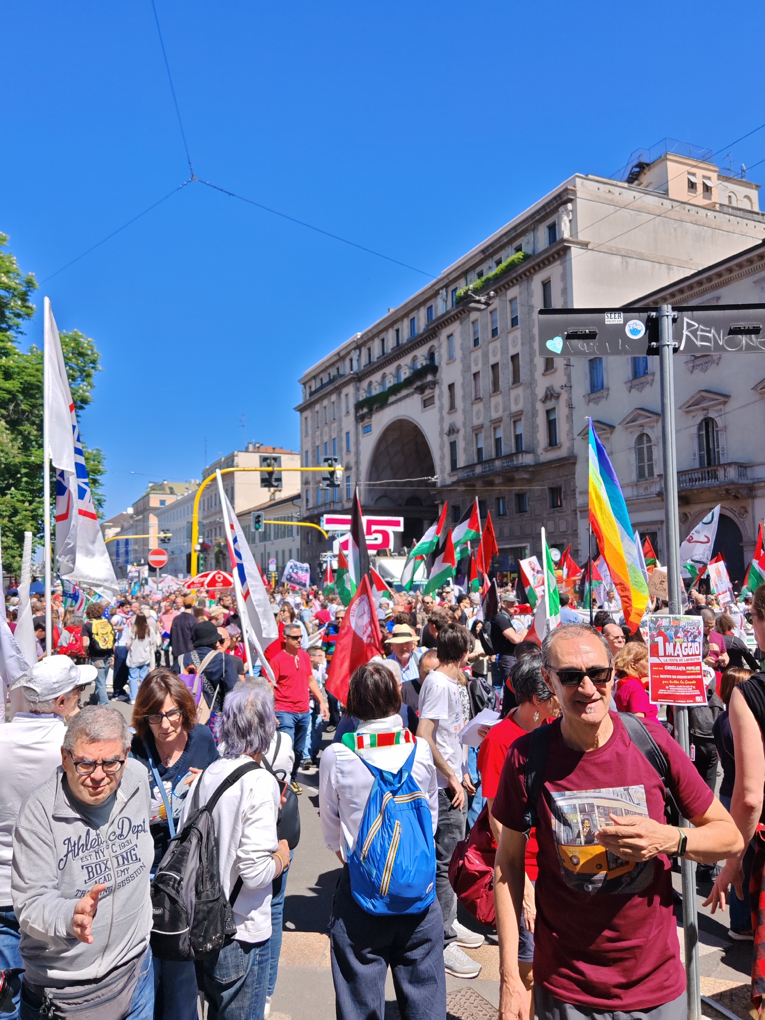 A large crowd of people fills a street in Milan, Italy, under a clear blue sky. They are gathered in front of a grand building with an arched entrance and numerous windows. Flags, including those of Palestine and rainbow flags, are visible amidst the crowd. Some individuals wear Italian flag scarves. Signs are held up, one specifically advertising "1 MAGGIO," a holiday celebrating laborers, with a flyer for a "GRIGLIATA POPOLARE," indicating a popular barbeque. A man in the foreground wears a burgundy shirt featuring a yellow tram. The crowd is varied, with individuals wearing casual clothing and backpacks. Street signs and traffic lights are also visible.
