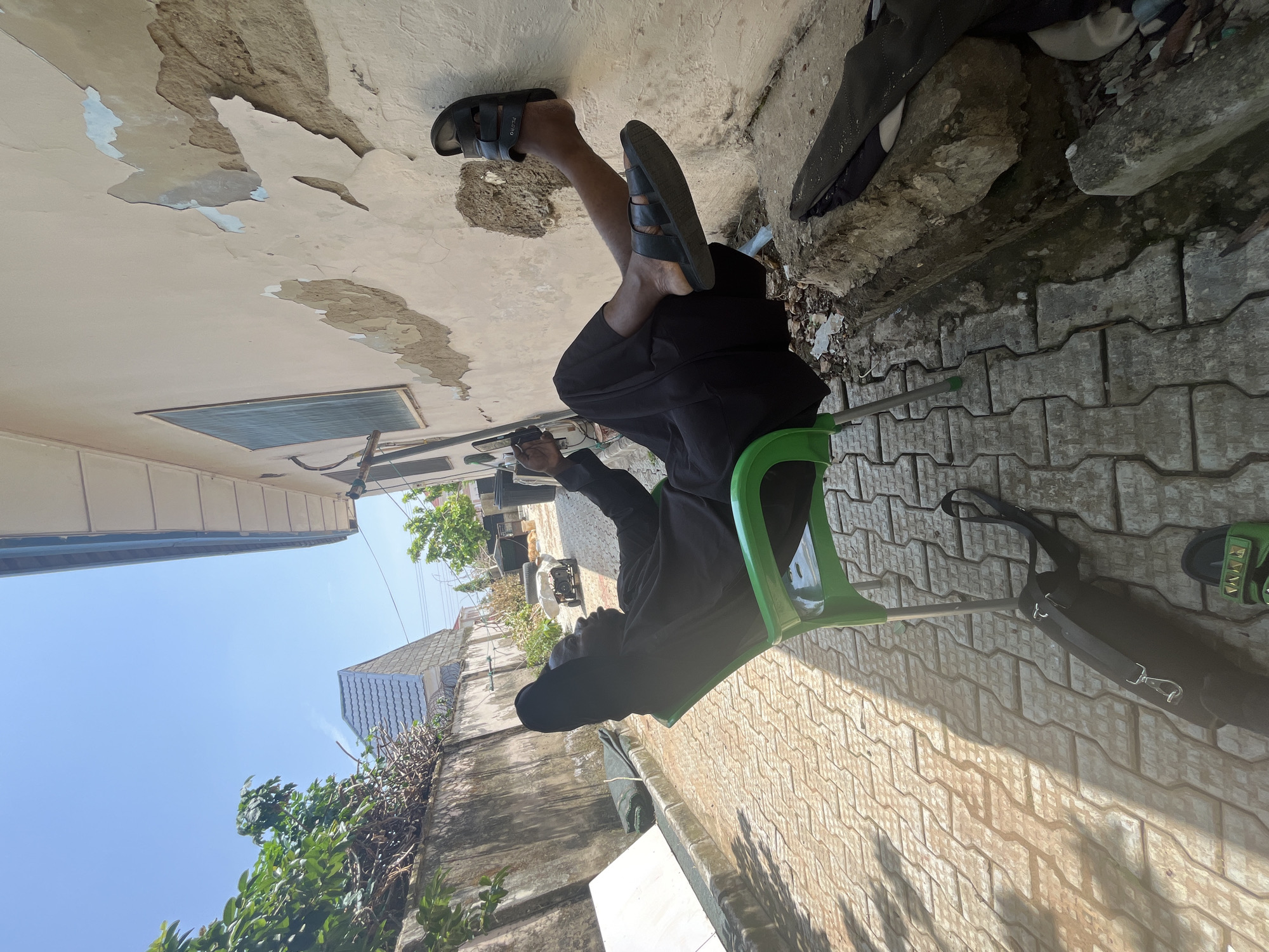 A man, dressed in black robes and sandals, sits on a green plastic chair outdoors, his legs propped up against a peeling stucco wall. The setting appears to be a narrow alleyway with interlocking brick paving. To his left, a black bag with a strap rests on the ground. Behind him, a concrete wall with foliage and a building with a tiled roof are visible. The sky is clear and blue. The scene is illuminated by bright sunlight, casting shadows on the ground. The overall impression is one of a casual moment in an urban environment, likely in Abuja, Nigeria, given the architectural style and general surroundings.