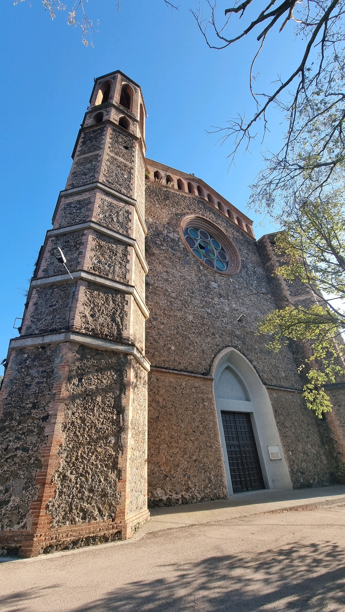 The image shows a low-angle shot of a stone and brick church with a prominent bell tower. The church has a circular stained-glass window above a large, dark, arched double door. The bell tower, constructed from stone and brick, has arched openings at its upper level. The exterior walls of the church are rough-hewn stone, contrasting with the brick accents. Bare tree branches are visible against a clear blue sky. The church is located in Sant Joan Les Fonts, Spain. The ground in front of the church is paved with asphalt, and shadows from the trees are cast across it.