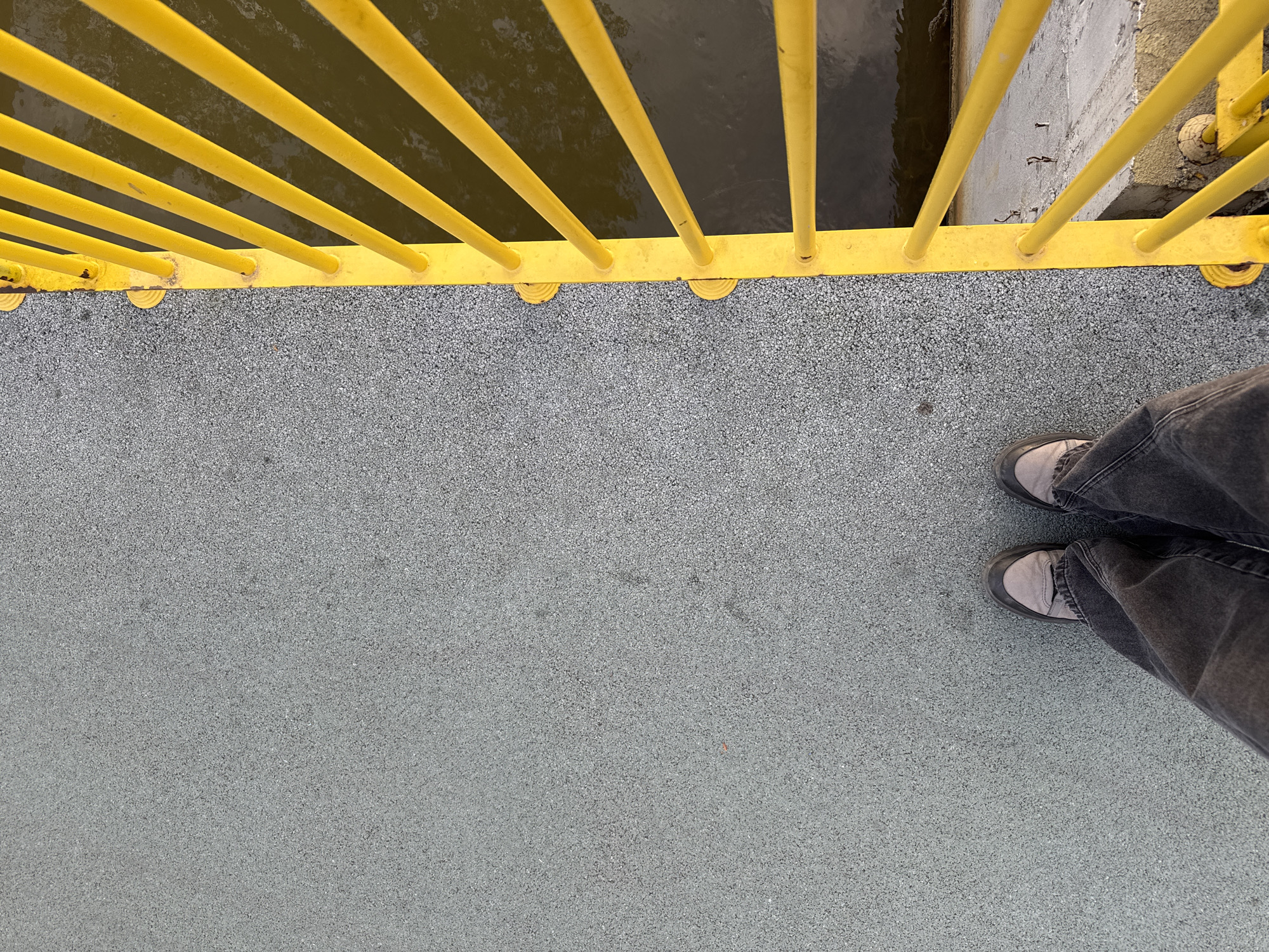 An overhead, eye-level shot captures a person standing on a textured grey surface next to a bright yellow metal railing. The railing, made of numerous vertical bars, runs horizontally across the top third of the frame, separating the pedestrian surface from a dark body of water below. On the far right, beyond the railing and partially submerged in the water, a concrete wall or support structure is visible.
The grey surface, resembling asphalt or a rubberized pathway, is granular and stretches across the foreground and midground. The person's lower legs and feet are positioned on the right side of the image, clad in dark, relaxed-fit jeans and light grey sneakers with dark soles.
The water appears dark and calm, possibly reflecting the sky or indicating depth. The yellow railing shows minor signs of wear and tear. The scene depicts an outdoor setting, likely a bridge, pier, or boardwalk overlooking a waterway, under daylight conditions. No specific time of day or weather conditions are discernible beyond general daylight. There is no visible text.