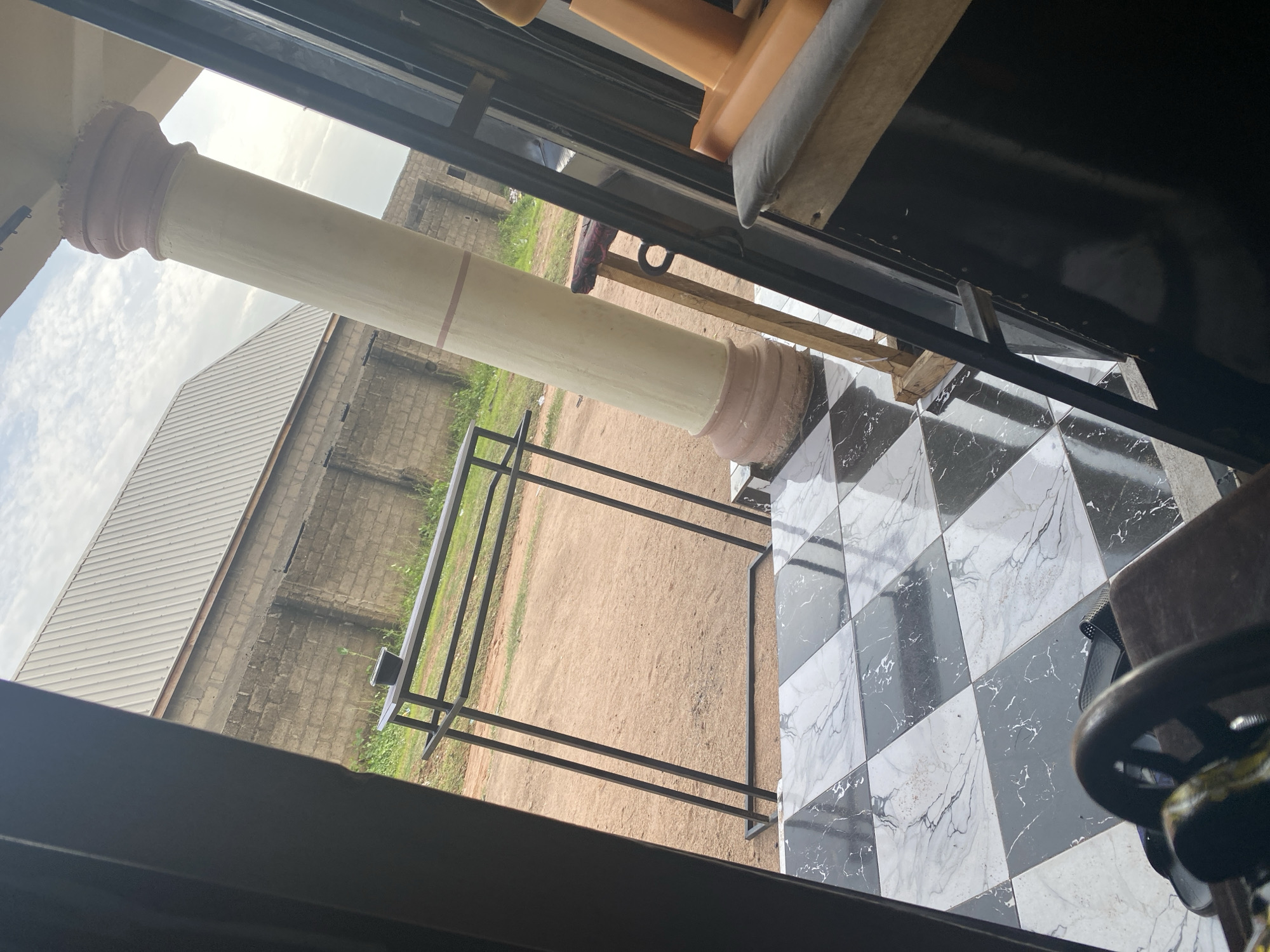 The image shows an outdoor scene, possibly a storefront or entryway, in Jalingo, Nigeria. A black metal clothing rack stands on a checkered black and white tiled floor. In the background, a building constructed of grey bricks with a corrugated metal roof is visible. A large, cream-colored pillar with a reddish-brown base is also present, supporting an overhead structure. To the right, a part of an orange plastic chair and a dark surface, possibly a table or counter, are visible. The ground behind the rack appears to be dirt. The sky is overcast with white clouds.