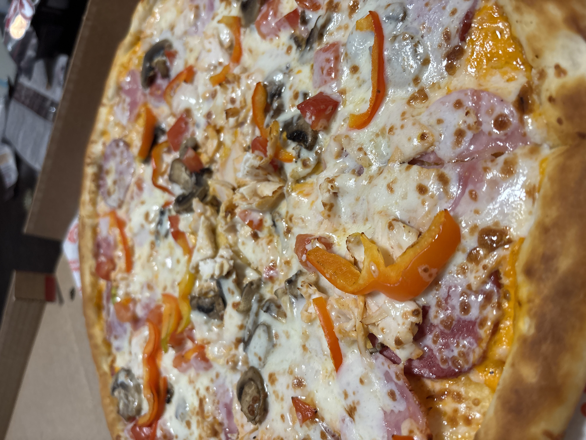 A close-up, high-angle shot shows a whole pizza with various toppings in a pizza box. The pizza is topped with melted cheese, sliced tomatoes, mushrooms, salami, bell peppers, and what appears to be chicken. The pizza crust is golden brown and slightly raised. The pizza box is cardboard with a brown exterior and a white interior, with some red and black printing visible on the lid in the background. The lighting is artificial, casting a warm glow on the pizza. The image is taken from a domestic setting, likely a home or restaurant, in Zhukovo, Russia.