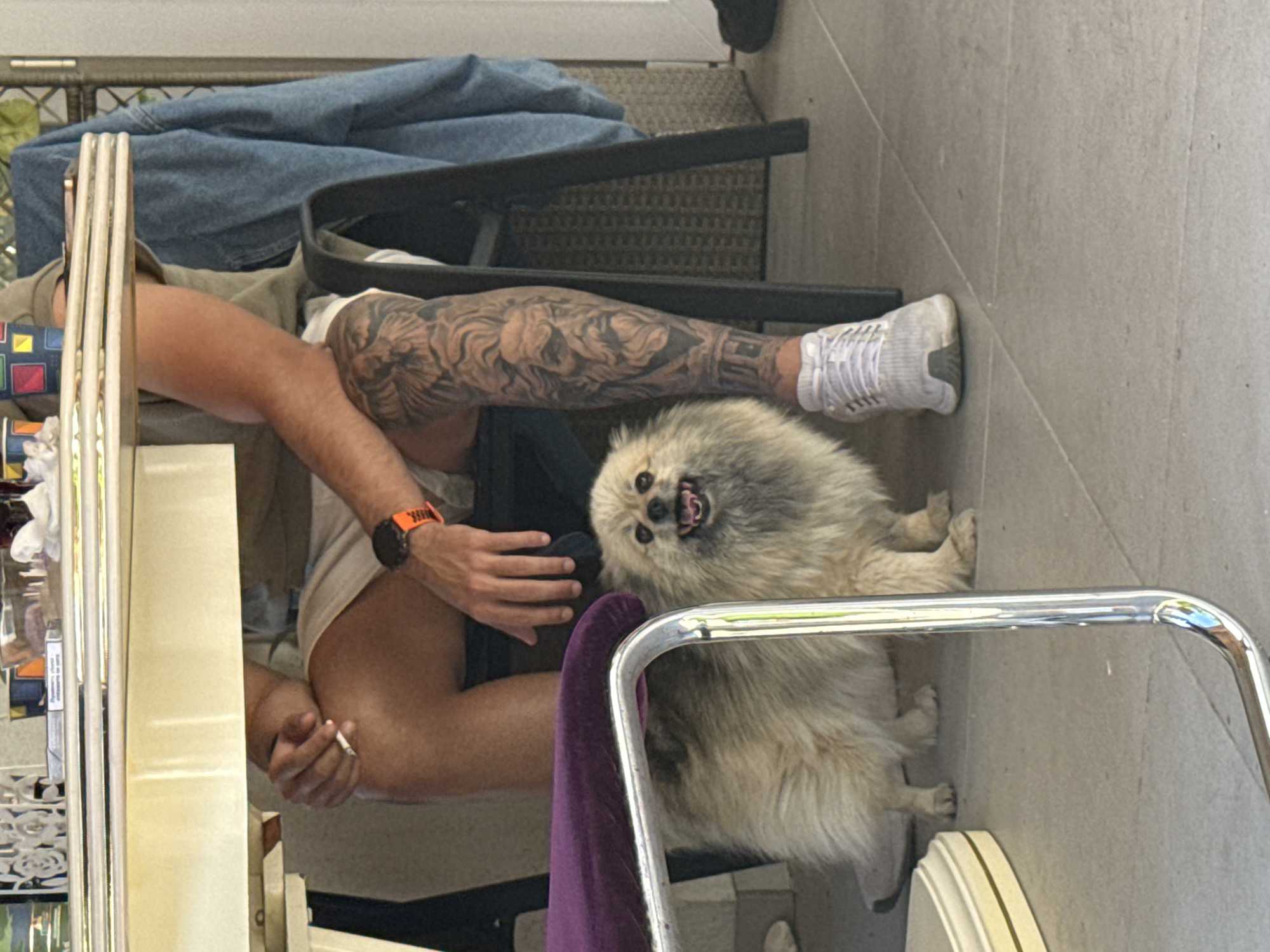 Outside in Elhovo, Bulgaria, a man sits at a table, his right leg with a detailed tattoo extending toward a dog. The dog is tan and white, and appears to be smiling, facing upwards. The man wears a light-colored shirt, black shorts, an orange sports watch, and white shoes. He is holding a cigarette. A blue denim jacket is draped over the back of the chair next to him. The table is covered with items. The scene is sunlit.
