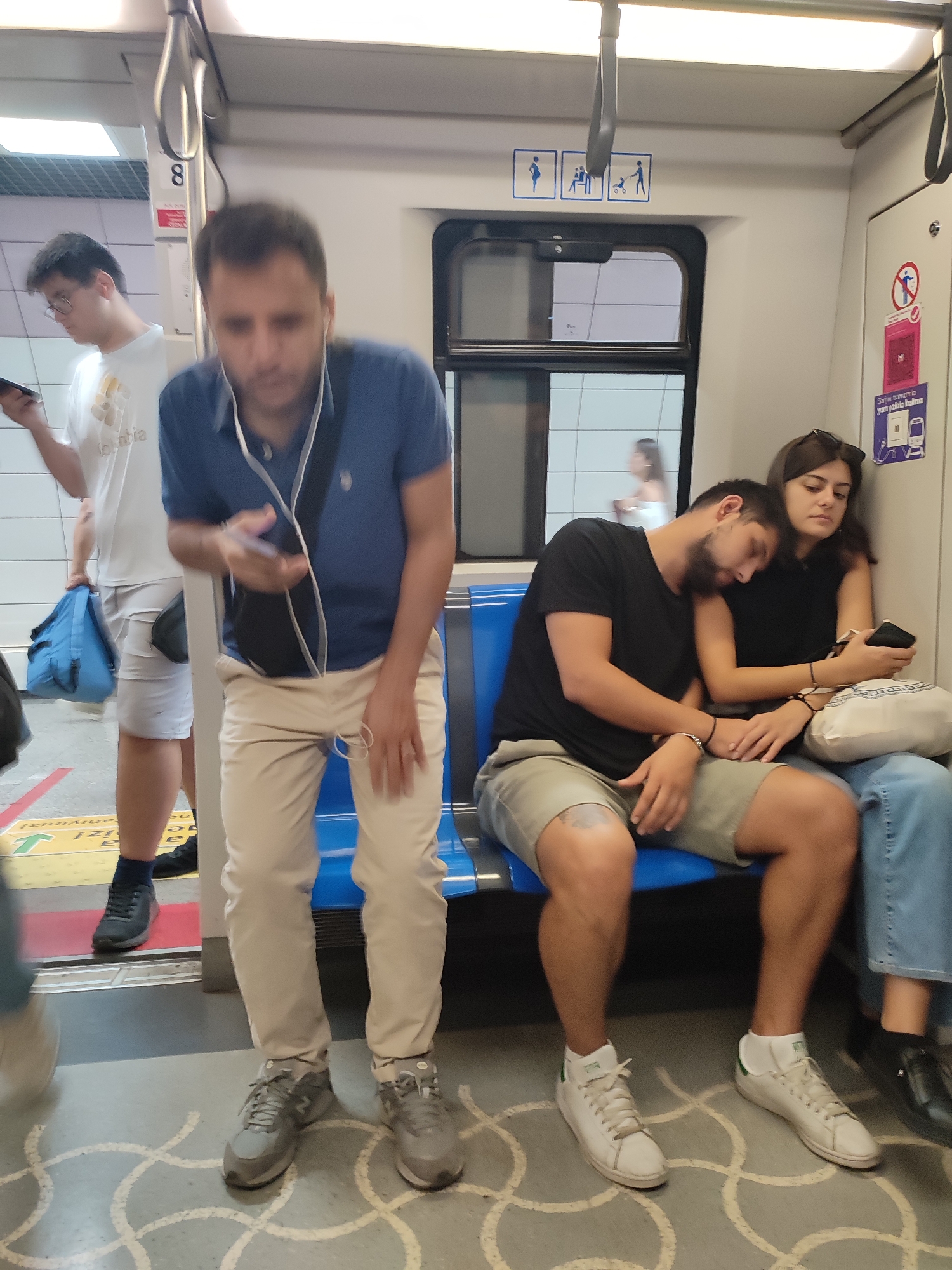 The image depicts the interior of a commuter train in Üsküdar, Turkey. In the foreground, a man in a blue polo shirt and light-colored pants stands, holding a phone and wearing earbuds. To his right, a couple is seated on blue upholstery. The man, with a beard, rests his head on the woman's shoulder. The woman, with dark hair, holds a phone and wears a black top. Further to the left, a young man in a white t-shirt and light shorts stands, holding a tablet, with a blue bag at his feet. The train car has visible grab handles, safety signs, and a window showing an out-of-focus background. The floor is grey with a white circular pattern.