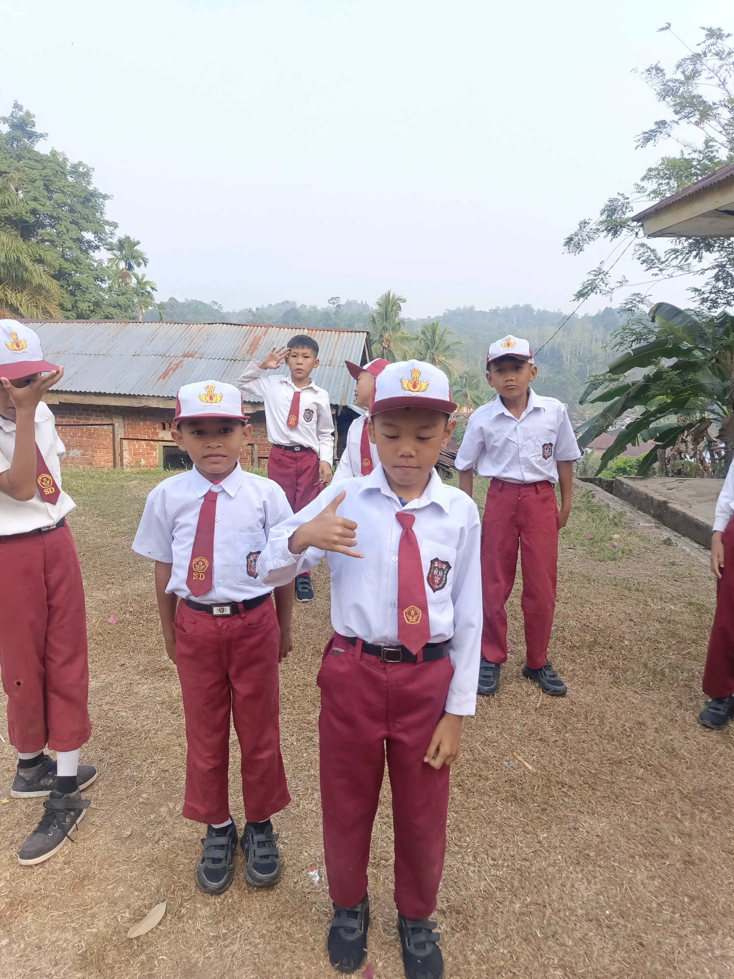 In Taba Tinggi, Indonesia, a group of young boys in school uniforms stand outdoors on a dry, grassy field. The boys are wearing white shirts with red ties, red pants, and white caps with red trim and a yellow emblem. Some of the boys are saluting, while others are looking at the camera or giving a thumbs-up. In the background, there is a weathered corrugated metal roof of a building, some trees, and a hazy sky. The scene appears to be a school event or gathering.
