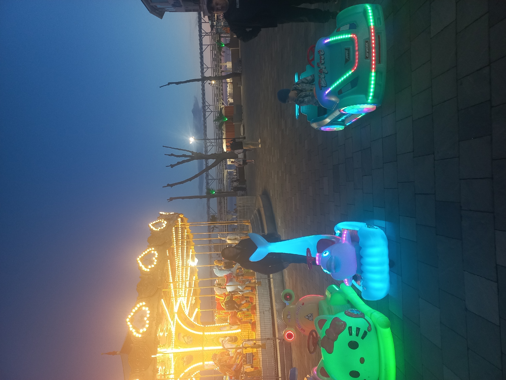 The image captures a vibrant night scene at an amusement area in Sochi, Russia, likely a boardwalk or pier. The time of day is evening or night, indicated by the dark blue sky and the predominant artificial lighting from rides and streetlights.
In the foreground, a child is seated in a glowing light blue ride-on car with LED lights in green and red. The car has "Bigfoot" written on its side, along with "Matrix" and "JD WORK" in smaller text. An adult stands behind this car. Adjacent to it, another brightly lit blue animal-shaped ride (possibly a dolphin) is visible with a person on it. A green Hello Kitty-themed ride is partially visible in the bottom right, also illuminated.
In the mid-ground, a brightly lit carousel with numerous yellow lights, including heart-shaped designs, dominates the left side of the frame. Several individuals, likely children, are visible on the carousel animals. Another person stands near the carousel's fence, which has "ВЫХОД" (Exit in Russian) signage.
The background features bare trees with streetlights and a structural framework, possibly part of a pier or an elevated walkway, stretching horizontally across the scene. The ground is paved with what appears to be dark tiles. The overall atmosphere suggests a lively leisure environment.