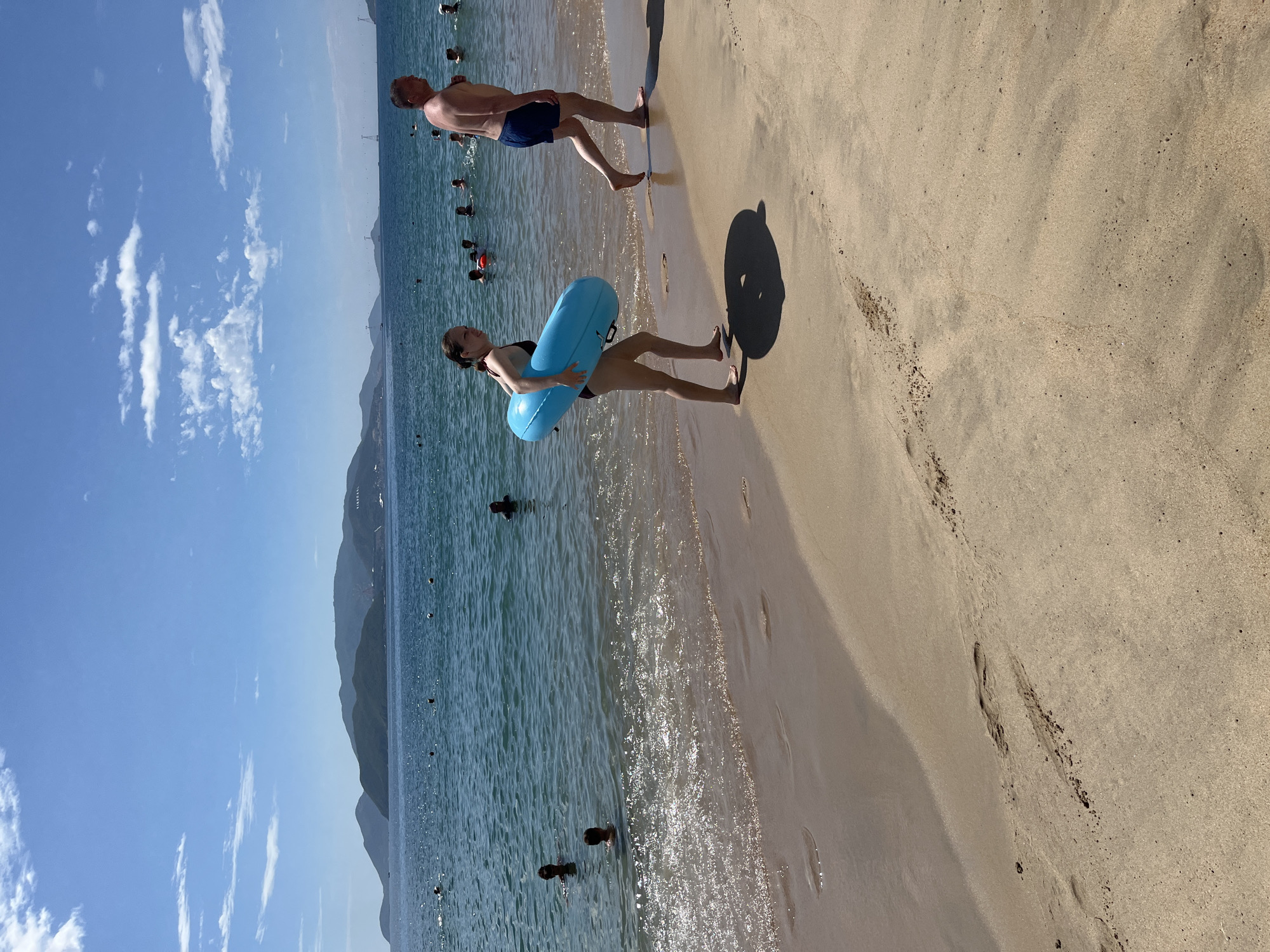 A bright, sunny day at a sandy beach in Nha Trang, Vietnam. The sea is calm and blue, with small waves lapping at the shore. In the foreground, a young woman with dark hair, wearing a black bikini, walks along the wet sand carrying a large, inflatable blue ring. Behind her, a man with a shaved head, wearing blue swim trunks, walks in the same direction. Both are walking away from the camera. The beach is dotted with other people swimming and wading in the ocean. In the background, a chain of green, mountainous islands stretches across the horizon under a clear blue sky with scattered white clouds.