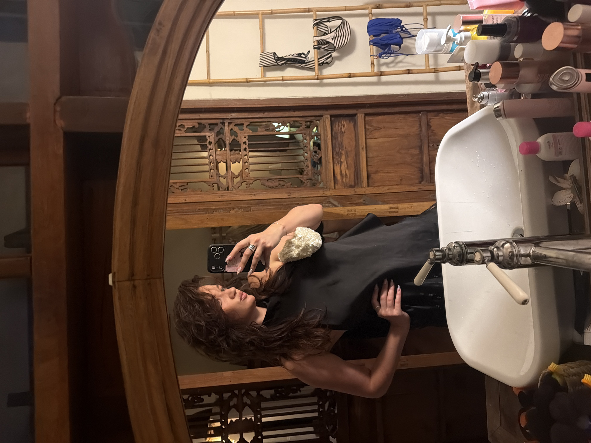 A woman is captured in a mirror selfie within what appears to be a bathroom or dressing area, characterized by its traditional wooden decor. She has long, wavy dark hair and is wearing a dark, possibly black, top or dress. She holds a smartphone to take the picture, her face visible in the reflection.
The large, arch-topped mirror she is using features a substantial, intricately carved wooden frame. Reflected in the mirror, to the woman's right, is a white porcelain sink equipped with vintage-style chrome faucets and cream-colored handles. Above the sink, a shelf holds numerous bottles of toiletries and cosmetic products.
The background visible in the reflection showcases ornate wooden paneling and carvings, indicative of traditional craftsmanship. Higher up on the wall, a bamboo rack is mounted, from which striped hair accessories and blue fabric items are hanging. The indoor setting is illuminated by warm, artificial light, suggesting evening or low-light conditions. The detailed wooden elements and bamboo accents throughout the room provide strong location cues pointing to a Southeast Asian, likely Indonesian, aesthetic, aligning with the provided context of North Kuta, Indonesia. The woman's expression is neutral as she poses. No specific text is clearly discernible on any of the items or surfaces.