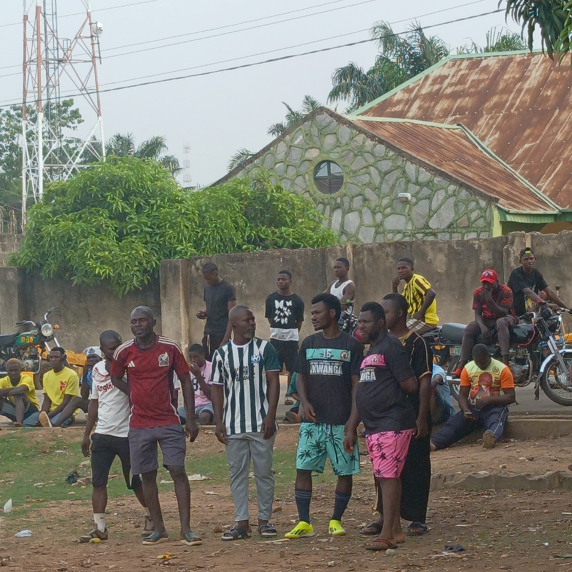 This image captures a casual gathering of men and young men outdoors in Akwanga, Nigeria, under an overcast sky. The scene is set on a dirt ground with sparse patches of grass and some litter.
In the foreground and midground, a diverse group of men stands and sits. Many are dressed in casual t-shirts and shorts. Visible text on their clothing includes "England" on a white shirt, "OPED FOOTBALL ACADEMY AKWANGA" and "UGA" on black shirts, and "MWANGA" on a pair of black and blue shorts. One man wears bright yellow football boots, while others have sandals or sneakers. Some are observing, some appear to be conversing, creating a relaxed, social atmosphere. Several motorbikes are present; one is ridden by a man on the right, and another with the number "1730" on its plate is partially visible on the left.
Behind the gathering is a concrete wall, and further back, lush green trees. A distinct building is visible on the right with a stone-patterned facade featuring a unique round window. Its roof is made of heavily rusted corrugated iron sheets. A tall, prominent telecommunications tower dominates the left side of the background, with another fainter tower visible in the distance. The overall impression is that of a community space, possibly a roadside or open area, where people gather.