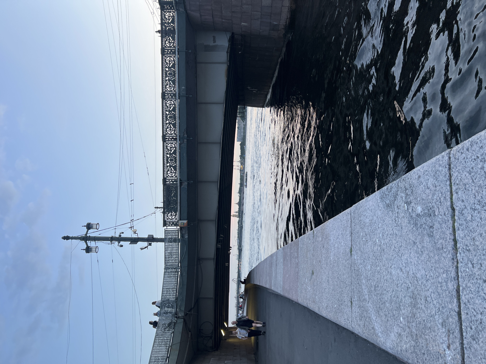 A wide, slightly elevated shot captures a scene along a waterfront in an unknown city. The water, a deep, dark blue, occupies the right two-thirds of the frame, reflecting the soft, diffused light of what appears to be dusk or early morning. Gentle ripples disturb the surface. A concrete embankment, composed of large, gray, rectangular slabs, lines the left edge of the water. To the left of the embankment is a paved walkway.
On the walkway, two individuals are walking away from the camera. The person on the left wears a light-colored top and dark pants. The person on the right wears a dark top and shorts. Further ahead on the walkway, more people are visible, some standing and some walking.
Above the walkway and embankment is a large bridge. The underside of the bridge is dark, with structural elements visible. The bridge's railing, made of ornate dark metal, is visible along the top. A tall, dark lamppost with two lamps stands on the bridge, near the left edge of the frame. Several wires are strung across the sky above the bridge and lamppost. In the distance, across the water, city buildings and spires are faintly visible against the pale sky, along with a few boats on the water.