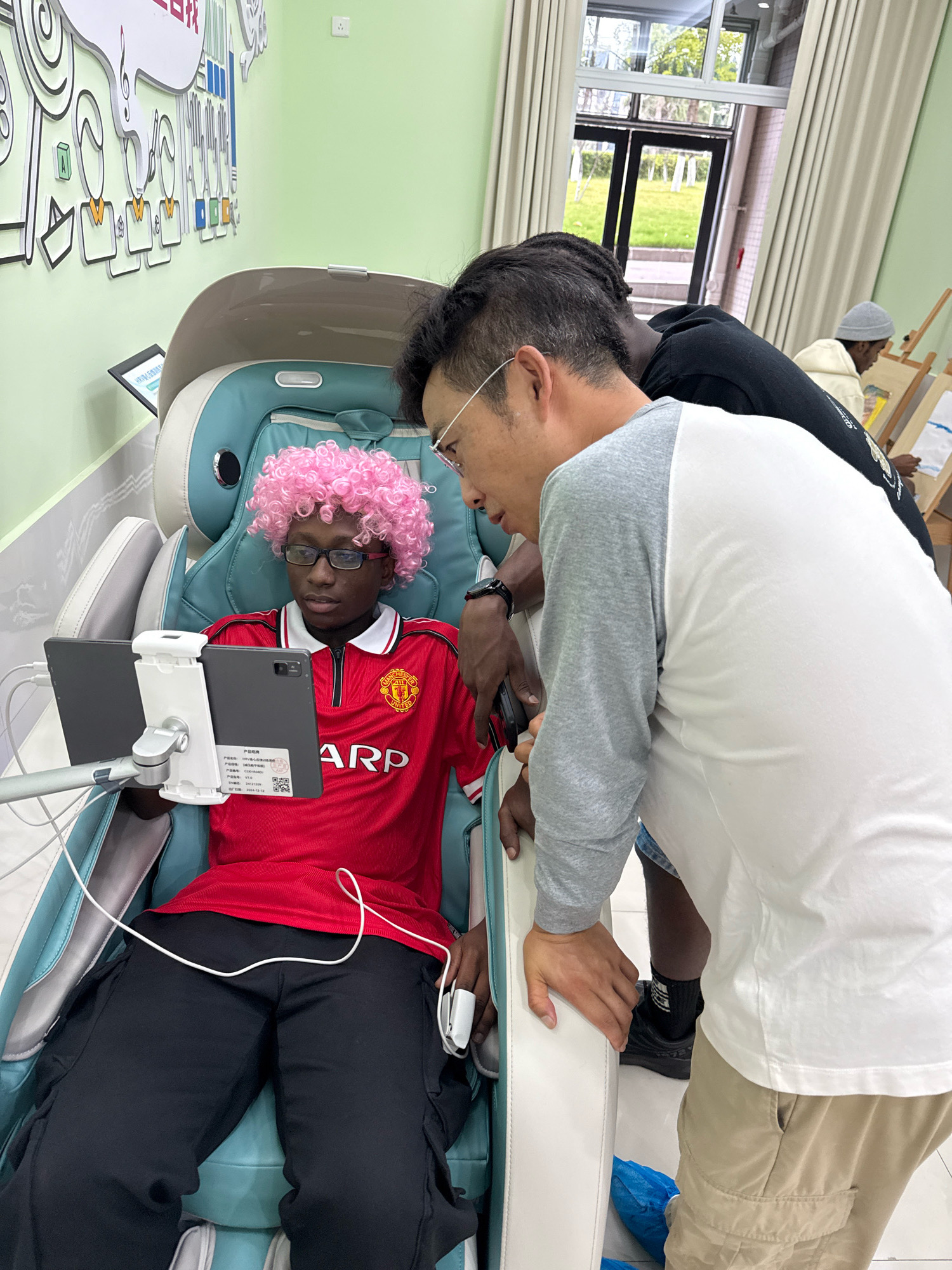In a brightly lit room with pale green walls, a young Black person wearing a red Manchester United jersey and a pink wig sits in a reclining massage chair, looking at a tablet. Two adult Asian men are leaning over the chair, one on the left with dark hair and glasses, and another on the right wearing a grey and white raglan shirt and a watch. The man on the right has his hand resting on the armrest of the chair, while the man on the left has his hands near the young person. A tablet is mounted on a stand in front of the young person, displaying some text. The room has a large window with beige curtains, revealing a glimpse of a grassy outdoor area. In the background, another person in a light-colored hoodie and grey beanie is seen standing at an easel, seemingly engaged in painting. A colorful mural with abstract figures and musical notes adorns the wall. The presence of art easels and the overall atmosphere suggest this could be a community center, art studio, or educational facility located in Jiangjin, China.
