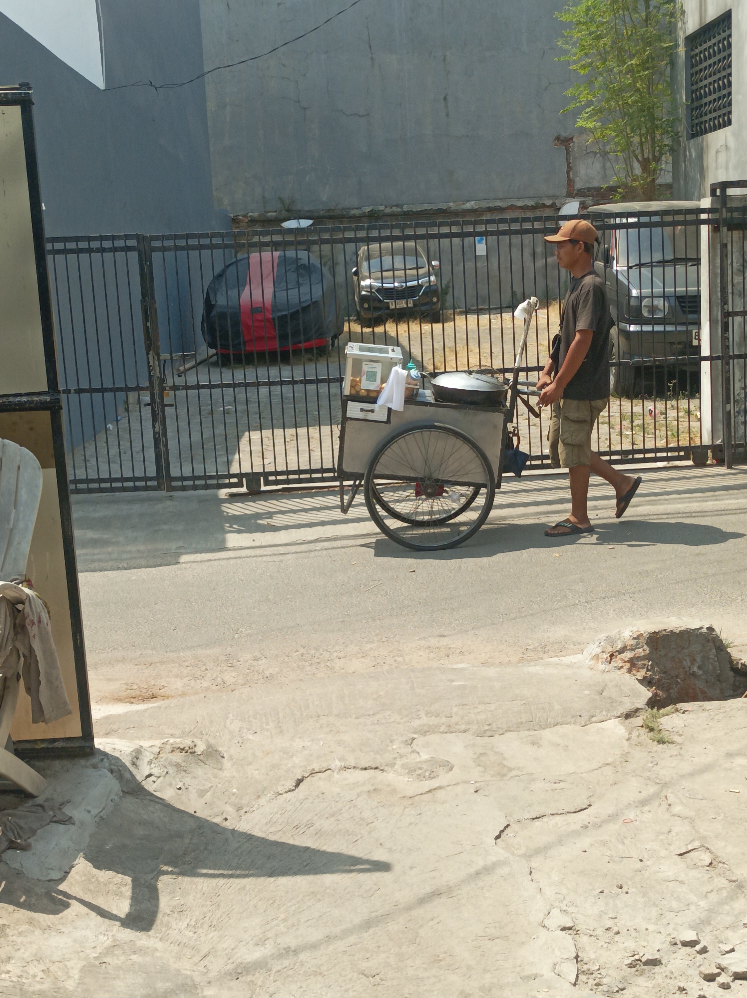 A man wearing a brown cap, a dark t-shirt, and khaki shorts walks with a food cart on a street in West Jakarta, Indonesia. The food cart is a metal structure with two wheels, a cooking pot, and various containers. He is pushing the cart to the right of the frame. In the background, there is a black metal fence, a gray concrete wall, and parked cars. Sunlight casts shadows on the street and sidewalk.