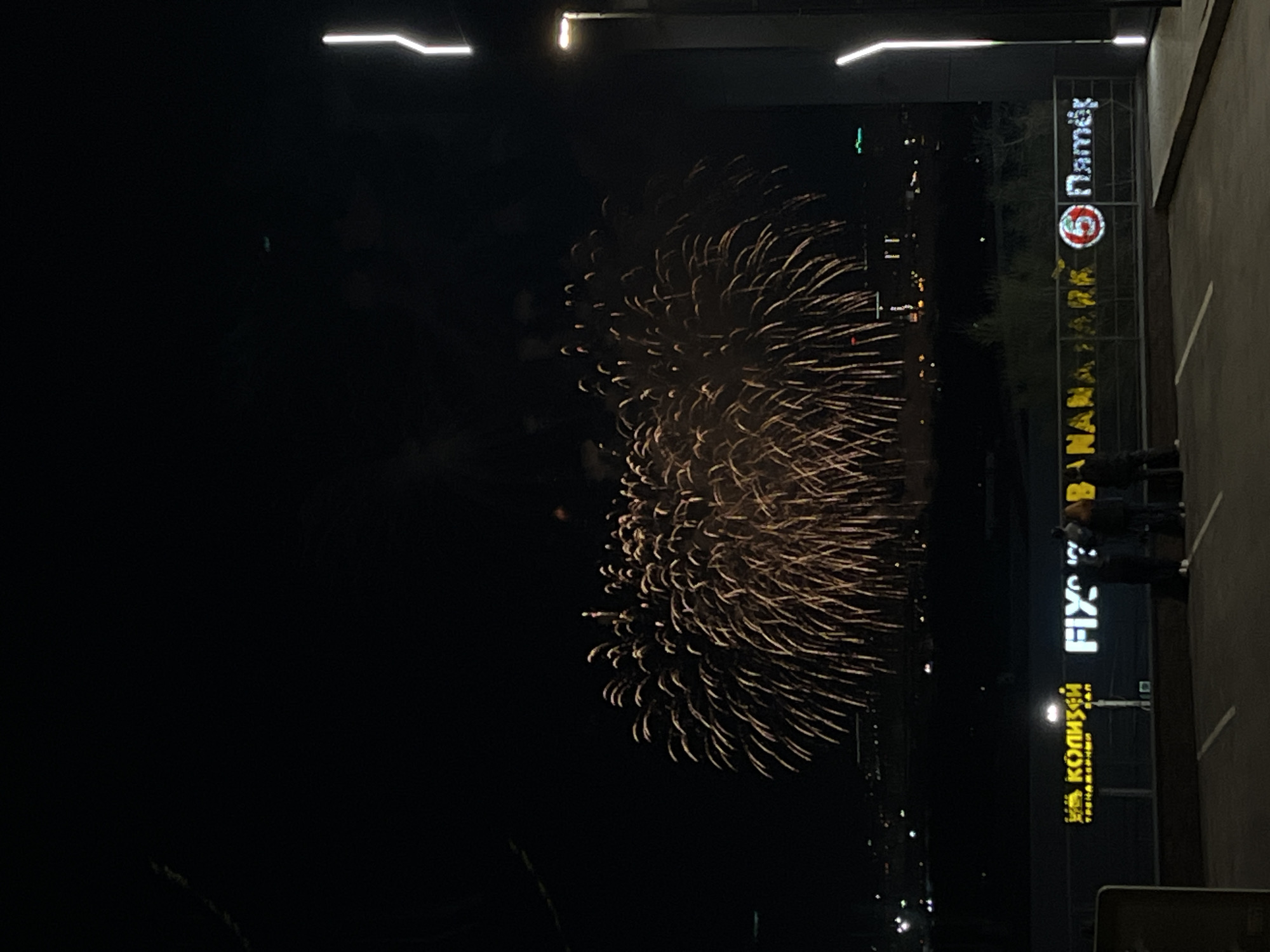 The image depicts a fireworks display in the night sky over Krasnoyarsk, Russia. The fireworks are bursting in a circular pattern, illuminating the dark surroundings. In the foreground, signs for "Пятерочка" (Pyaterochka), "Banana Park," "Fix," and "Колизей" (Coliseum) are visible, identifying the location. Several individuals are observing the fireworks from a nearby vantage point. The setting is urban, with streetlights and buildings discernible in the background.