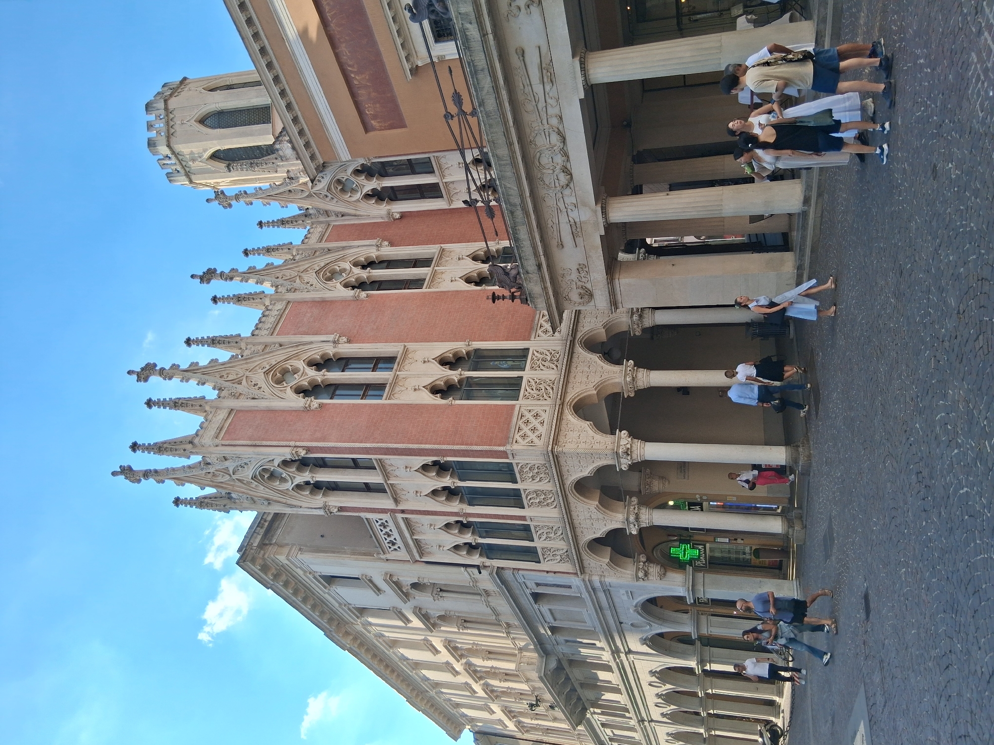 The image shows the exterior of the Palazzo Bo, University of Padua, in Padua, Italy. The building features a highly ornate Gothic facade with intricate stonework, pointed arches, and decorative pinnacles. A cylindrical tower with battlements rises above the main structure. Several people are walking on the cobblestone street in front of the building. Some are in groups, while others are walking individually. The sky is blue with a few scattered clouds. The scene depicts a sunny day in an urban environment with a historic architectural landmark.