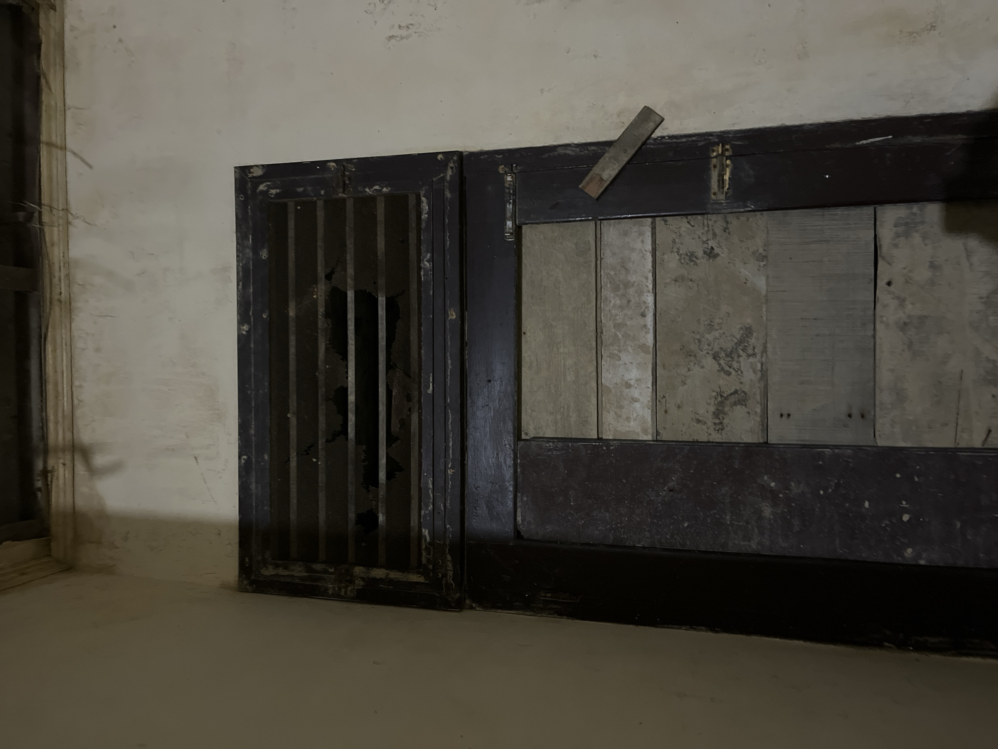 The image shows a dilapidated doorway in what appears to be an interior space. The doorway is constructed of dark wood and features a slatted vent at the top, some of which is broken. The lower portion of the door is made of planks of wood, some lighter and some darker, with visible wear and tear. A small, flat piece of wood is wedged into the frame of the door on the right side, suggesting a makeshift lock or support. The walls surrounding the doorway are discolored and show signs of age and neglect. The scene is dimly lit, contributing to the overall impression of disrepair. The location is specified as Kota Kuala Simpang, Indonesia.