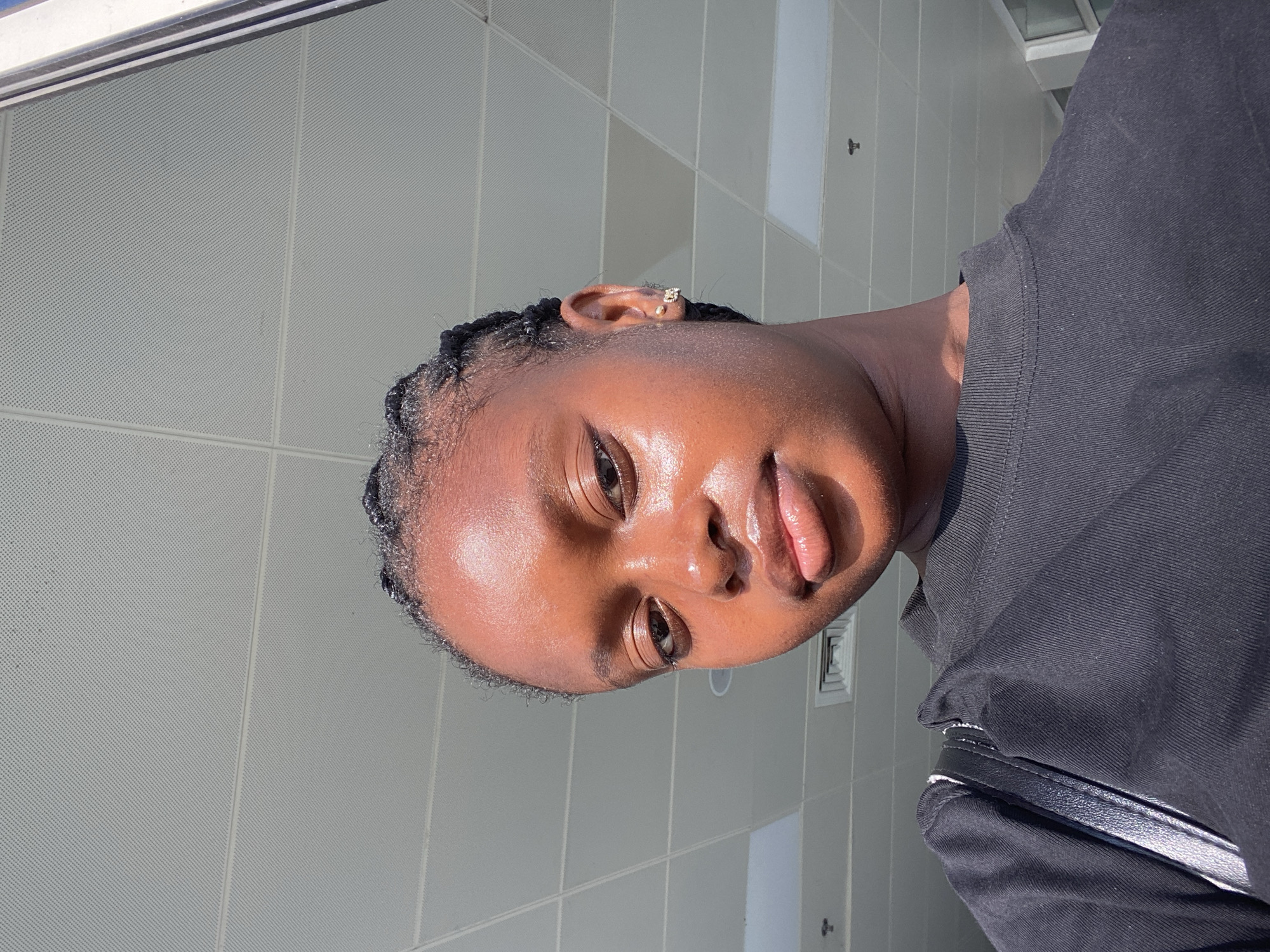 A person is angled towards the camera, with a focus on their face. They have dark skin, braided hair, and an earring in their left ear. They are wearing a dark-colored shirt. The background consists of a light gray wall with a grid-like pattern. A vent or small panel is visible on the wall. The location appears to be indoors. The specific city is unknown.