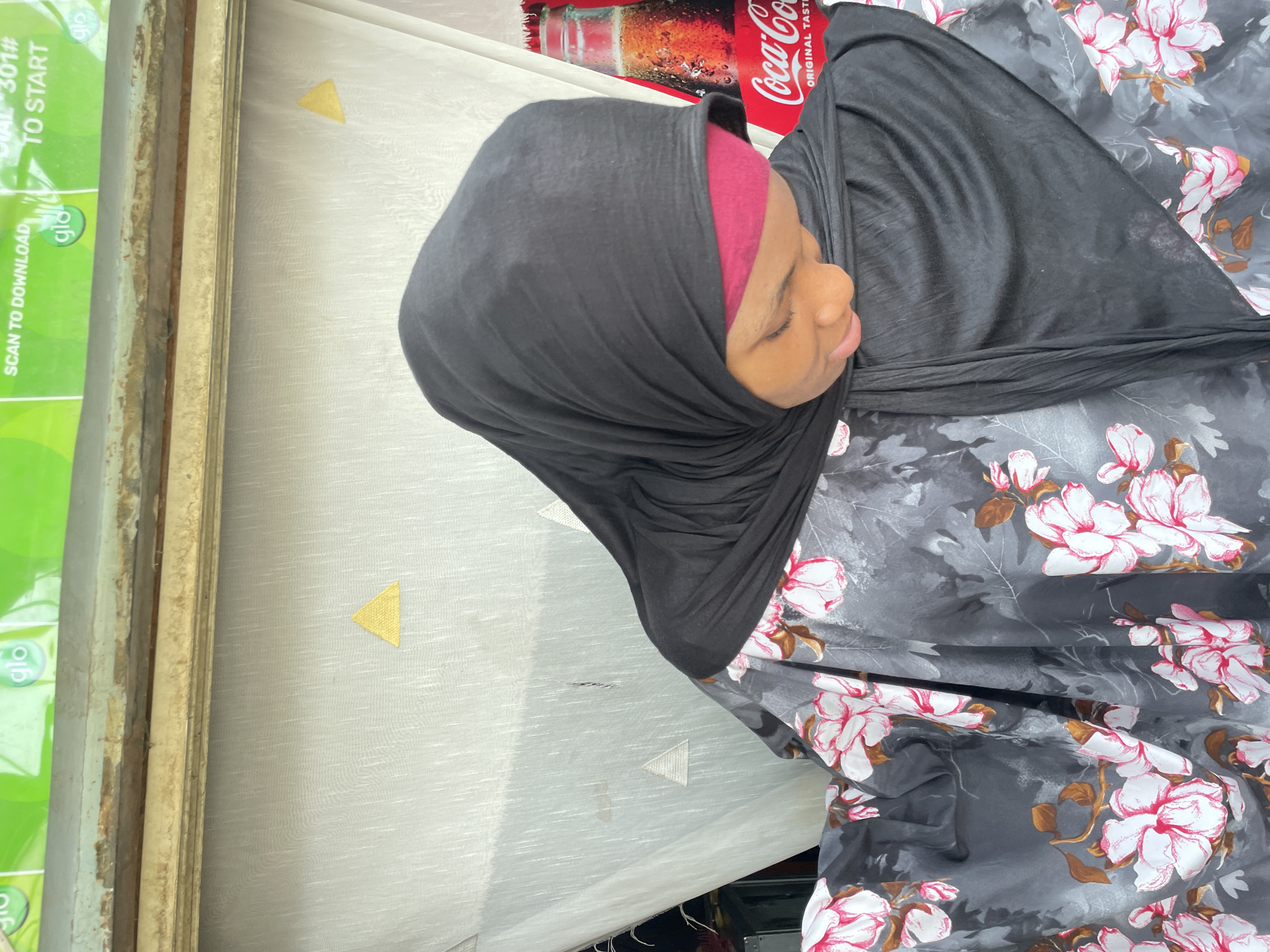 Here is a forensic description of the image.
A medium, canted shot presents a person in a black hijab with a burgundy underscarf. Adjacent to the person, there's a Coca-Cola product. In the background, one can see a window with a sticker. The sticker contains the words "glo", "SCAN TO DOWNLOAD", "AL 301#", and "TO START".