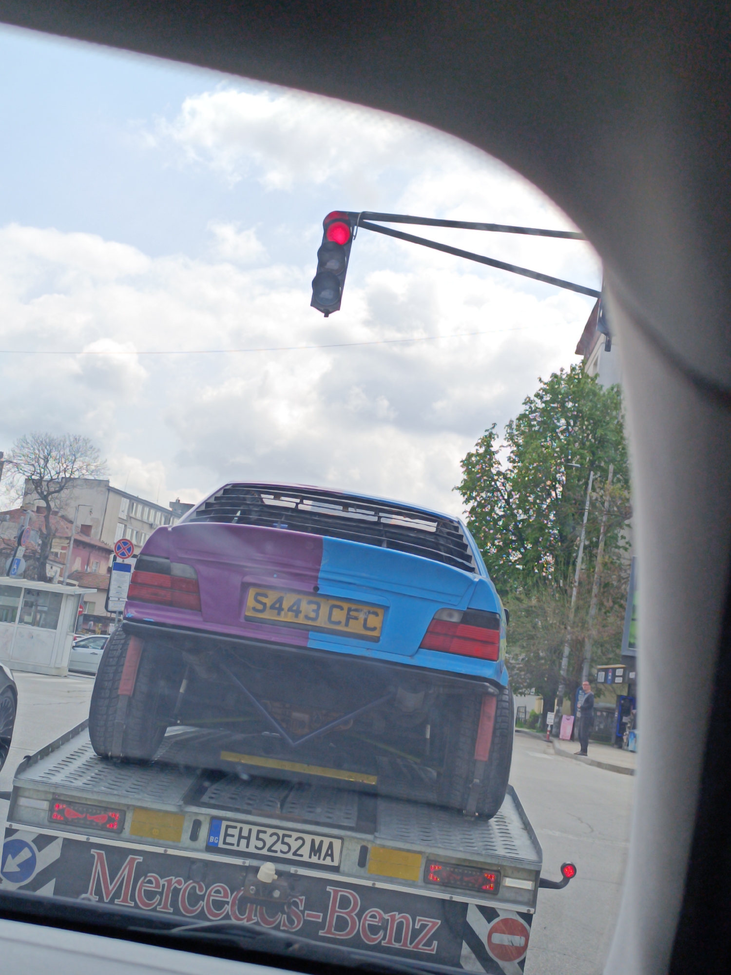 The image shows a two-tone, purple and blue car with a license plate reading "S443 CFC" being transported on a Mercedes-Benz tow truck with the license plate "EH5252 MA". The tow truck is in an urban setting. There's a red traffic light overhead. A person is standing on the sidewalk nearby. The presence of a bus stop structure and other buildings suggest the location is the city of Pleven, Bulgaria. The image appears to be taken from inside another vehicle.