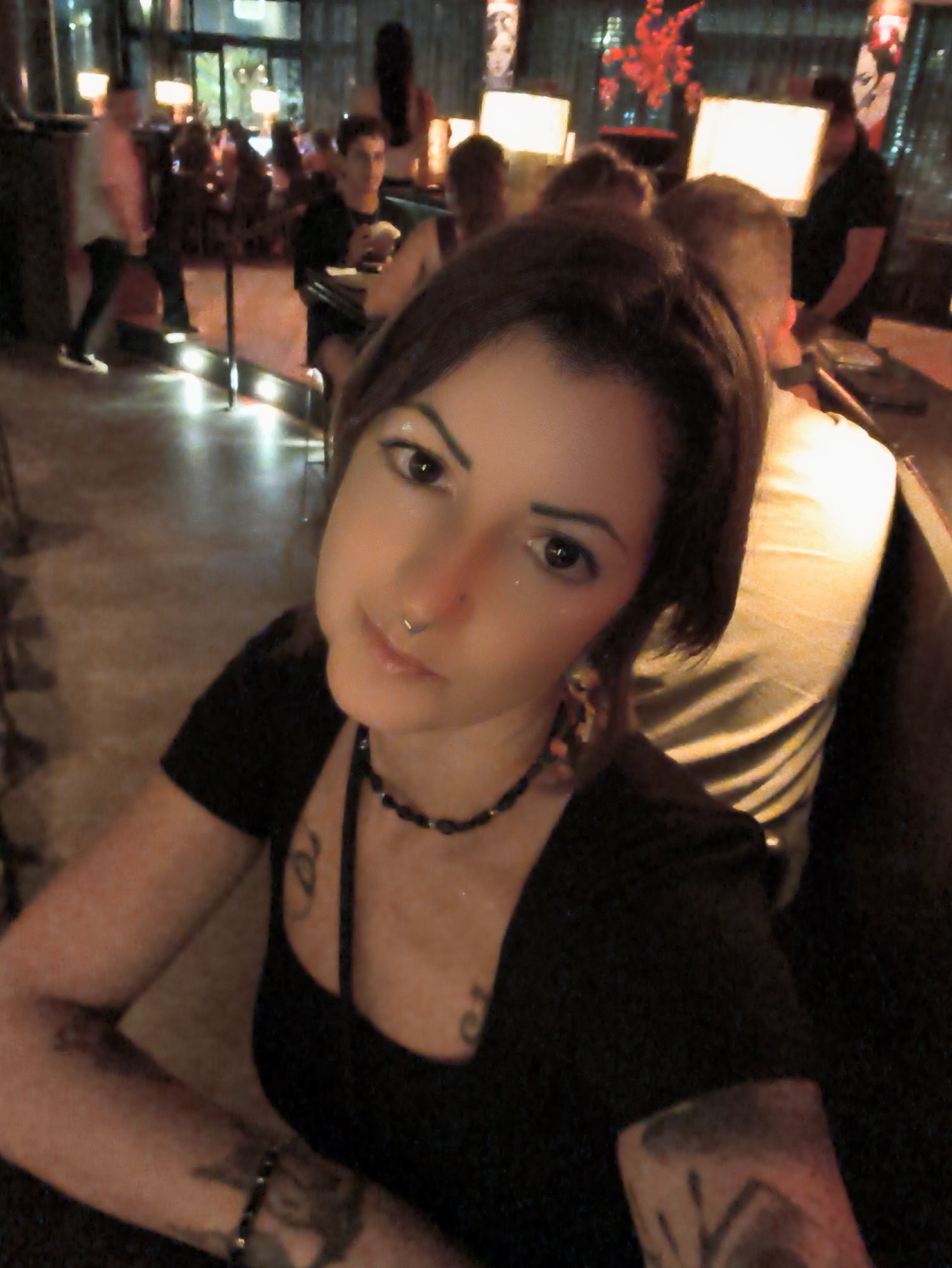 The image depicts a close-up of a woman with dark hair, brown eyes, and fair skin. She is wearing a black top and a beaded necklace. She has a nose ring and visible tattoos on her arms and chest. The background is blurred but suggests an indoor setting, possibly a bar or restaurant, with other patrons and lighting fixtures visible. There is no indication of the specific location in Be'er Sheva, Israel.