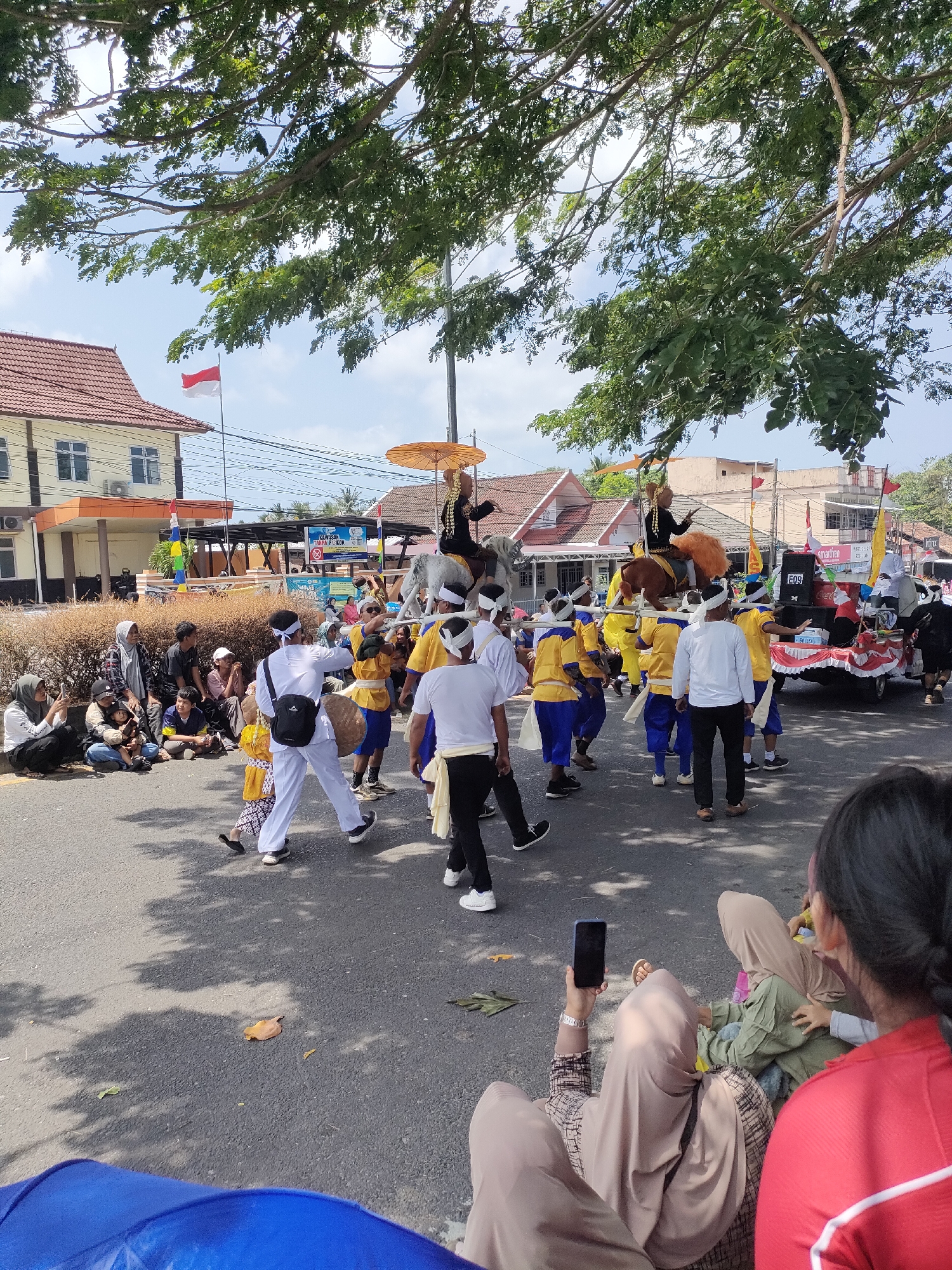 This image depicts a street parade or festival in Sungailiat, Indonesia. A group of performers in traditional attire, including white headbands and yellow and blue outfits, are participating. Some are carrying a decorative float or effigy that appears to be made of a horse-like structure. Two individuals are seated on horseback-like figures, holding golden umbrellas. Spectators are gathered along the side of the road, some sitting on the ground, observing the procession. A building with a tiled roof and several windows is visible in the background, along with trees and a bright sky. The street is paved, and shadows from trees are cast upon it. The presence of an Indonesian flag suggests a national or local celebration.