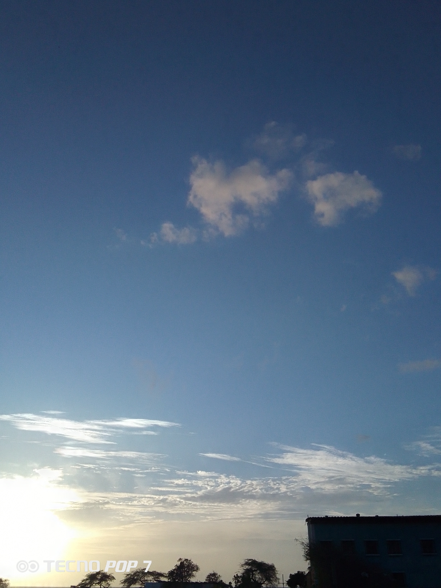 The image shows a daytime scene in Pedro González, Venezuela, focusing on the sky above the ground. The sky is predominantly blue with several white clouds scattered across the frame. At the bottom of the frame, there is a view of the horizon where the sun appears to be setting or rising. The sun's light illuminates the clouds, creating a bright and diffused glow. Below the horizon, there are trees and what looks like the silhouette of a building. The lower left corner of the image has a device logo with "TECNOP P7" visible.