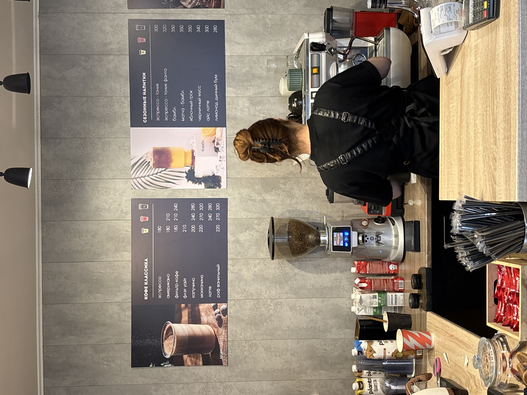 The image shows the counter of a coffee shop with a barista standing behind it. The coffee shop appears to be located in Chaikovsky, Russia, judging by the Russian writing on the menus. The barista, a woman with her hair pulled back, is wearing a black uniform and an apron. She is facing the coffee machine, and her hands are obscured from view, implying she might be making coffee.
To the left of the barista are two menus attached to a gray wall. The first menu is titled "Кофе Классика" (Coffee Classics) and lists different coffee drinks and their prices. The second menu is titled "Сезонные напитки" (Seasonal Drinks) and lists other coffee drinks and their prices.
On the counter, to the right of the barista, are a variety of items, including a coffee grinder filled with beans, boxes of milk, cups, straws, candy bars, a cash register, and other coffee-making equipment.