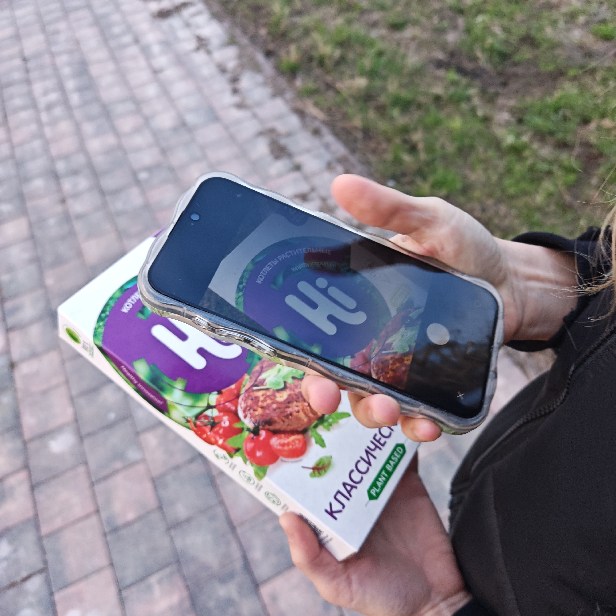 A person is holding a box of plant-based "Hi" brand classic burgers in their left hand, while holding a smartphone in their right hand, using the phone’s camera to view and possibly photograph the box. The box is white with purple lettering reading "КЛАССИЧЕСКИЕ" (Classic) and "PLANT BASED" in green. The "Hi" logo is displayed prominently on the box. The phone screen displays a camera interface and mirrors the "Hi" logo from the box. The background shows a sidewalk paved with brick tiles and some green grass and foliage, suggesting an outdoor location. Given that "Hi" is a Rostov brand, it is plausible this photo was taken in Rostov, Russia.