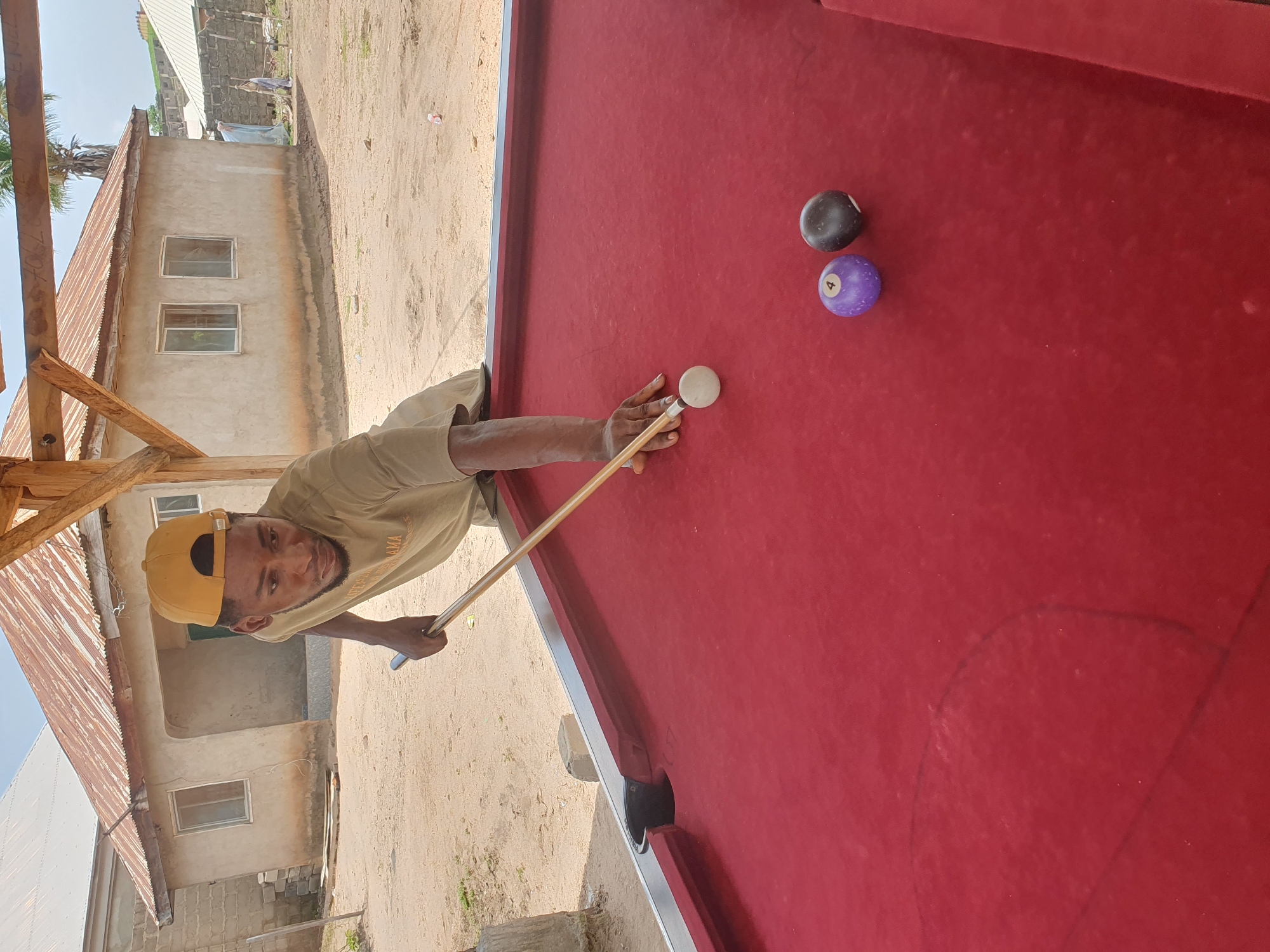 A man wearing a yellow baseball cap and a light brown t-shirt is playing billiards outdoors. He is in the process of striking the white cue ball with a cue stick. Two other billiard balls are visible on the red felt surface of the table: a purple ball with the number 4 on it, and a solid black ball. The billiard table is positioned under a wooden shelter, with a sandy dirt ground surrounding it. In the background, there are simple, one-story buildings with corrugated metal roofs, characteristic of a rural or semi-urban setting. The architecture suggests a location in Lokoja, Nigeria, with its typical building styles and outdoor living environments.