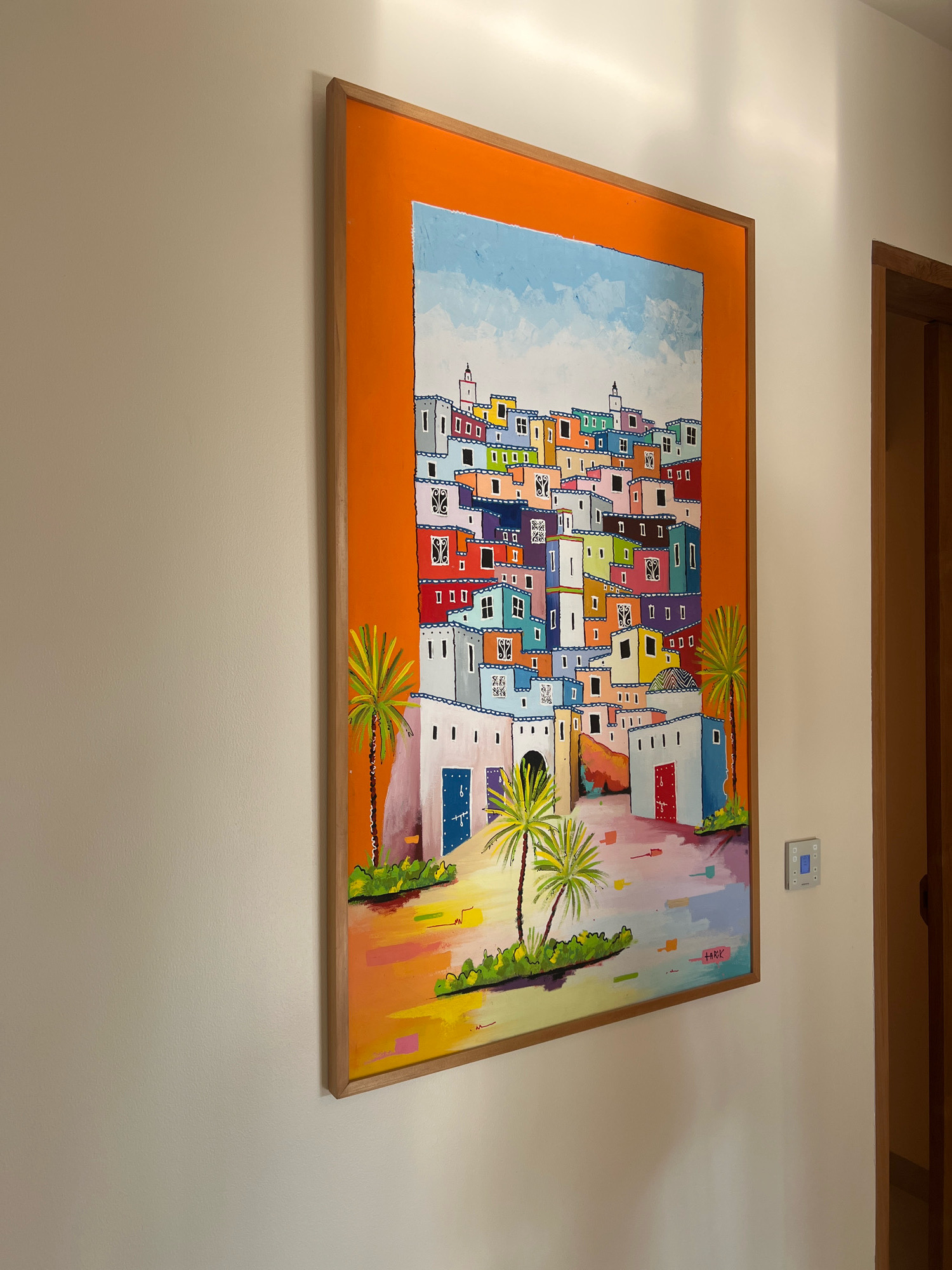 A brightly colored, folk-art painting of a coastal town is displayed on a cream-colored wall. The painting, framed in light wood, depicts a densely packed cluster of houses with red-tiled roofs and vibrant walls in hues of orange, yellow, green, blue, and purple. Palm trees with stylized fronds are visible in the foreground. The painting is signed with a red signature on the bottom right. To the right of the painting, a wooden door frame is visible, and a light switch panel is mounted on the wall. There is no indication of this location being in Ngaglik, Indonesia.