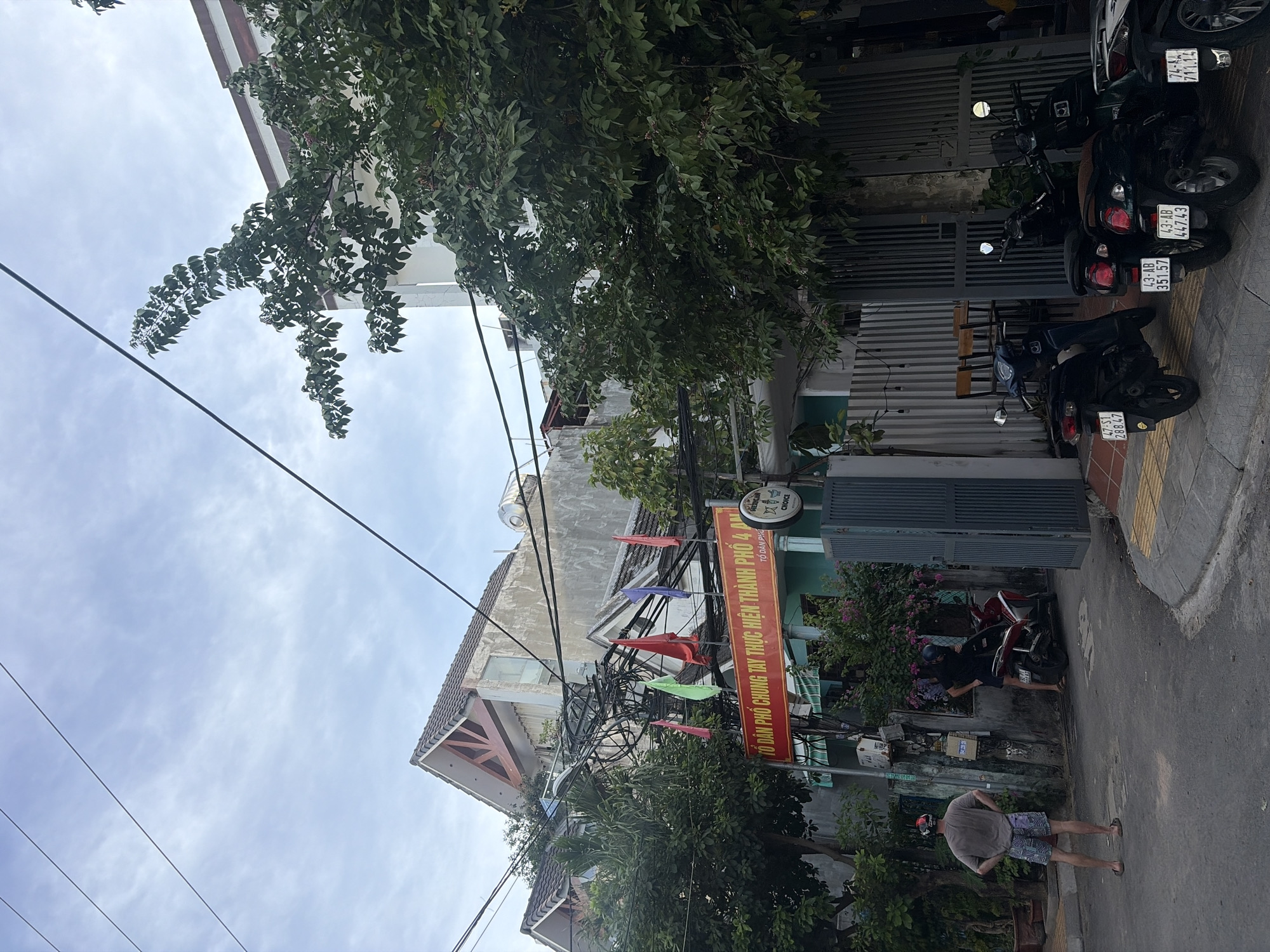 The image depicts a street scene in Đà Nẵng, Vietnam, with a clear view of residential buildings and parked motorcycles. A man in a grey t-shirt and patterned shorts stands facing away from the camera, his hands on his hips, on the left side of the street. He wears a helmet. To his right, another man in a black shirt and shorts is interacting with a red and black motorcycle. Several motorcycles are parked along the sidewalk on the right side of the street. A large banner with Vietnamese text hangs across the buildings, accompanied by several colorful flags. Power lines are prominently strung across the upper portion of the image, creating a visually complex overhead environment. The buildings appear to be a mix of residential and commercial establishments, with some featuring balconies and tiled roofs. The overall atmosphere is that of a typical urban street in Vietnam, with a blend of everyday life and architectural elements.