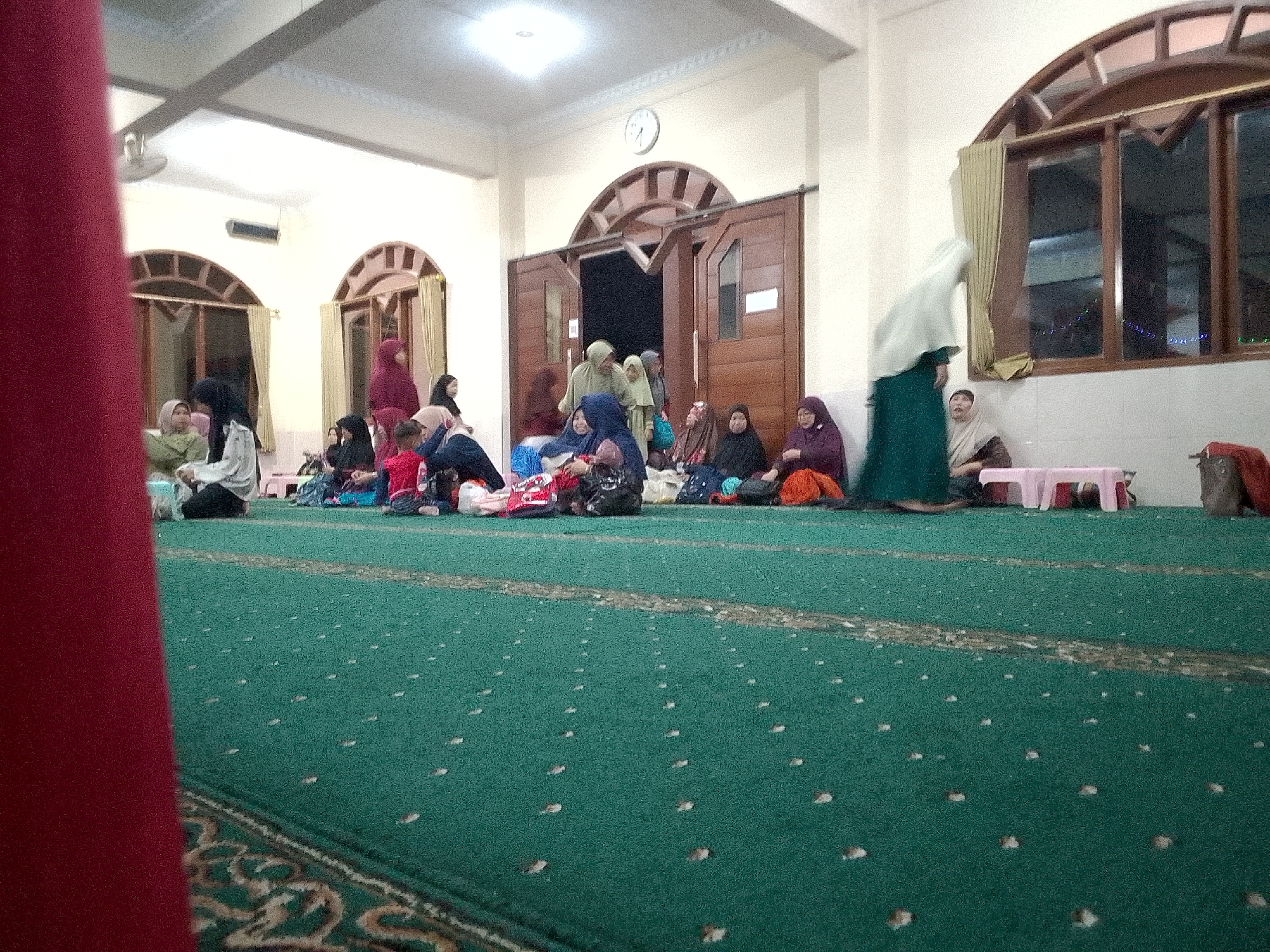 The image depicts an indoor scene, likely a mosque, with several people, predominantly women, seated on a green carpeted floor. The environment appears to be a communal gathering space, with architectural features such as arched doorways and windows visible. The individuals are dressed in modest clothing, with many wearing hijabs. Some are engaged in conversation or observing their surroundings, while others appear to be resting. Bags and personal belongings are scattered around the area, suggesting a prolonged stay or a transition period. The lighting suggests it is daytime. While the city of Kertajaya, Indonesia is mentioned in the prompt, there are no explicit visual cues within the image itself that confirm this specific location.