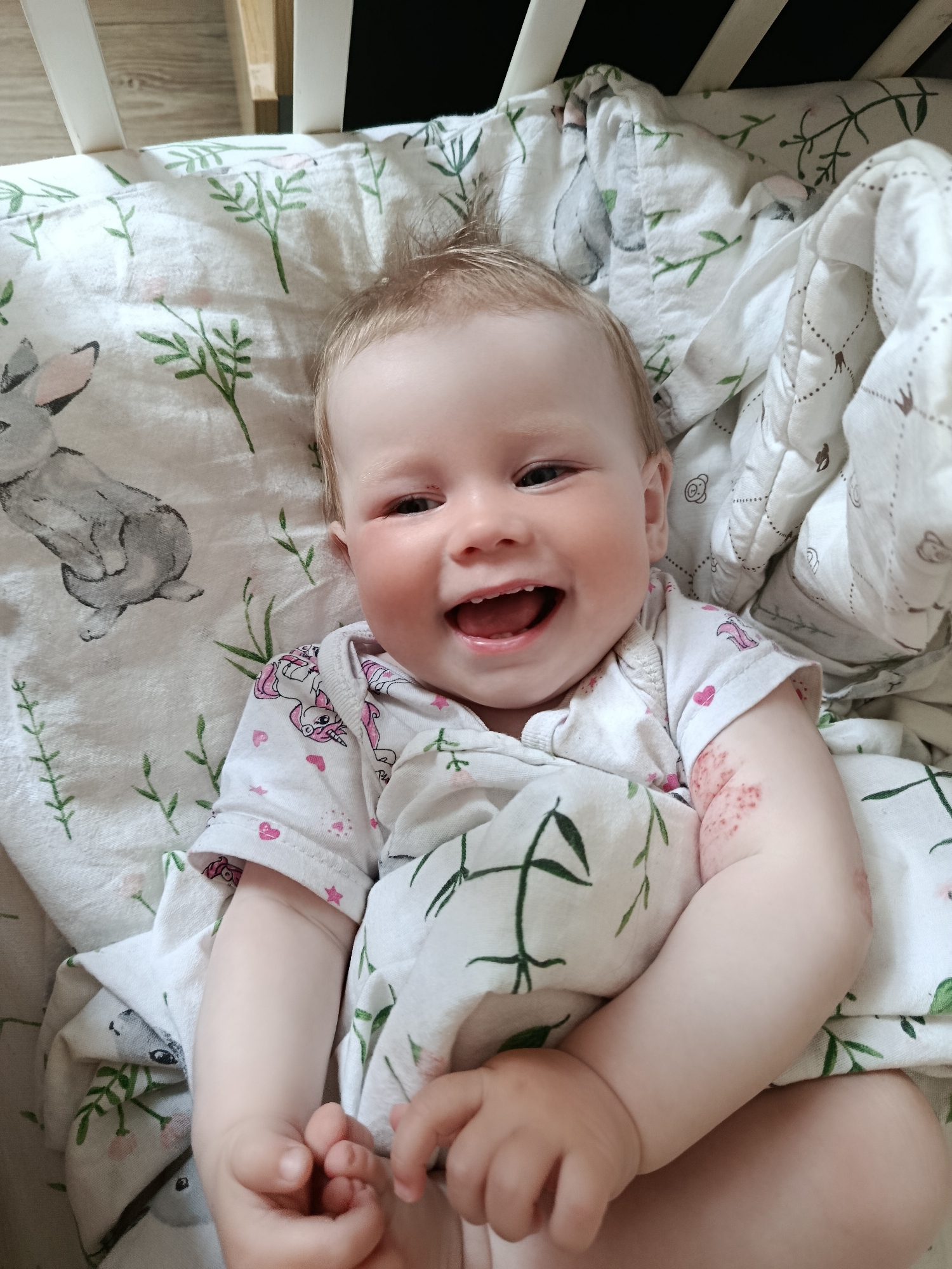 The image depicts a baby lying in a crib. The baby has fair skin, blonde hair, and is smiling widely, revealing some teeth. The baby is wearing a white onesie with a pattern of unicorns and hearts. The baby is wrapped in a white blanket with a pattern of green plants and a gray rabbit. The crib has white railings. The photo appears to have been taken indoors. The location is unknown.