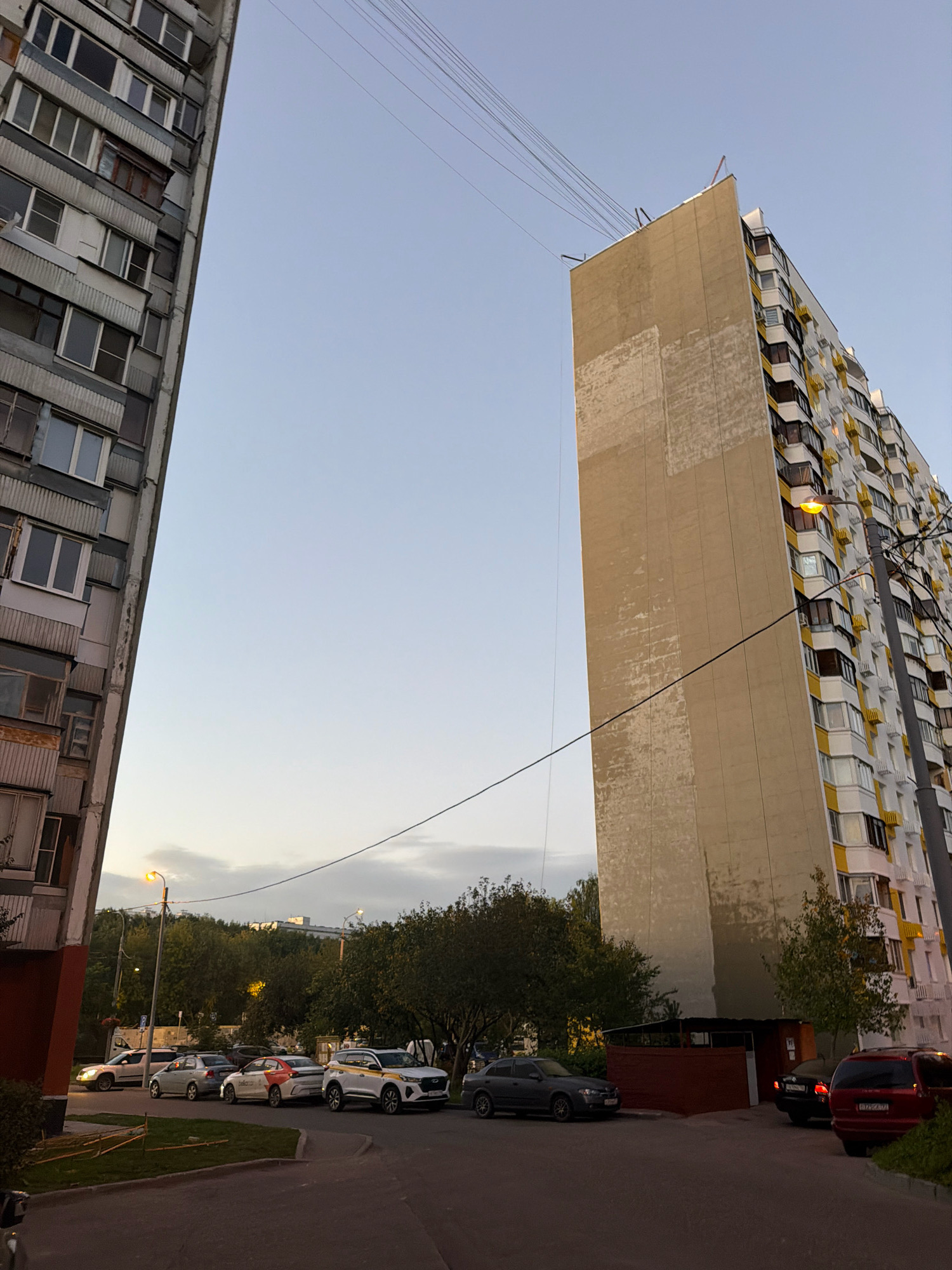 An urban scene in Moscow, Russia, captured from a low angle, features two multi-story apartment buildings dominating the frame. The building on the left is a typical Soviet-era panel building, its grey facade characterized by repetitive windows and balconies, some appearing weathered and showing signs of disrepair, including rusted metal elements. The building on the right is a more modern structure with a yellow and white exterior, featuring a multitude of balconies and windows, suggesting a residential complex. A large, pale beige-colored section of this building appears to be a blank wall, possibly an unfinished or unrenovated part. Overhead, a network of power lines stretches across the sky, connecting the buildings and contributing to the urban ambiance.
At ground level, a street is visible with several cars parked and driving. Among them, a white car with yellow and orange markings, possibly a taxi or ride-sharing vehicle, is prominent. Other vehicles include sedans and an SUV. The area around the buildings includes patches of grass and mature trees, particularly in the background, creating a contrast between the built environment and nature. Streetlights are visible, one casting a warm glow, indicating it is either dusk or dawn. The sky is a clear, pale blue, with a hint of cloud cover on the horizon. The overall impression is that of a residential area in a Russian city, likely a large urban development.