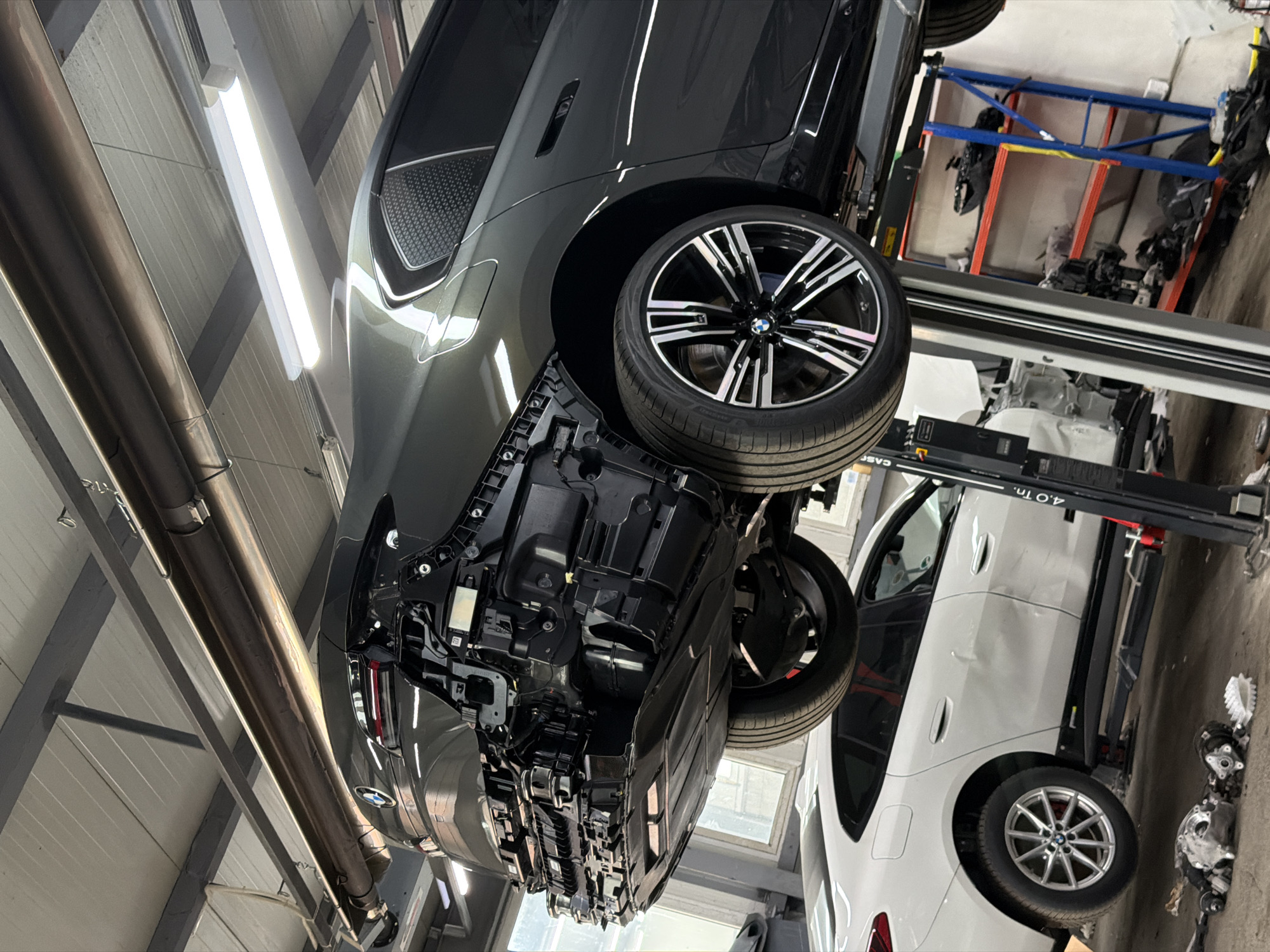 An overhead view within a car workshop in Burgas, Bulgaria, shows a dark grey BMW SUV, likely an iX model, elevated high on a car lift. The vehicle is seen from its rear-left side, with the rear bumper partially removed, exposing various underbody components, wiring, and structural elements. One of its large, multi-spoke alloy wheels with a BMW logo and a tire is prominent in the foreground.
Fluorescent tube lights illuminate the ceiling above, and large silver pipes run along the wall. In the background, part of a blue and orange storage rack with various parts is visible. Below the elevated car, partially obscured by the lift mechanism, a white BMW SUV (possibly an X3 or X5) is parked on the workshop floor, with its rear wheel and side visible.
The car lift itself is black and red, with visible text "4.0 T." indicating a 4.0-ton capacity, and a partially visible "CASE" or similar brand name on one of its supports. The scene suggests the dark grey BMW is undergoing service or repair work, with its undercarriage accessed for maintenance. The overall setting is a clean and well-lit automotive service bay.