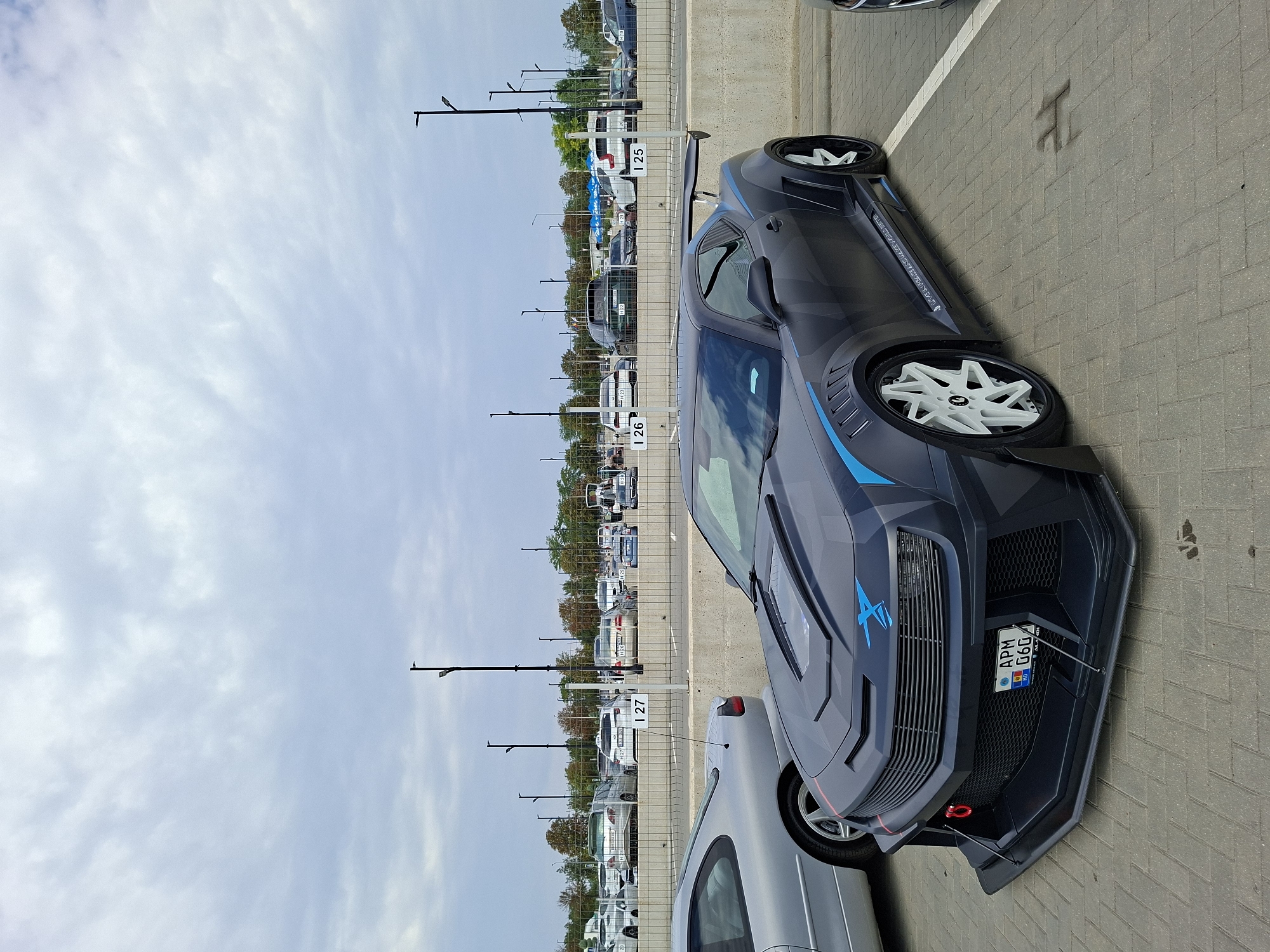 The image captures a modified dark gray Chevrolet Camaro parked in an outdoor parking lot. The car features an aggressive body kit, including a large rear spoiler, widebody fenders, and a custom front bumper with a red tow hook. The vehicle has a distinctive blue graphic design on the hood and fenders, and white multi-spoke rims. A license plate reading "APM G60" is visible. To the left, a silver coupe is partially visible. In the background, a concrete barrier separates the parking area from a row of other vehicles and trees. Streetlights are present in the background. The ground is paved with interlocking bricks. The sky is overcast with scattered clouds. The location is identified as Stăuceni, Moldova.
