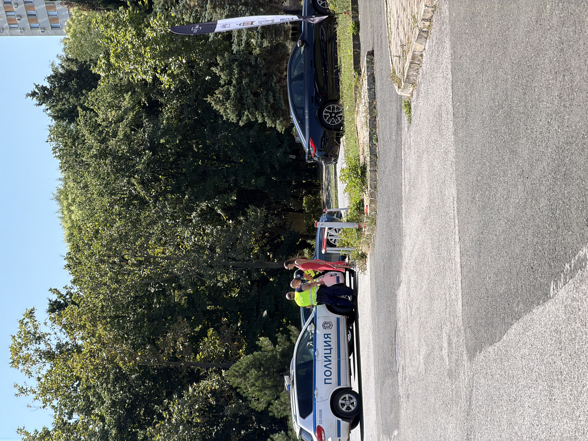 A police car is parked next to a black sedan in Burgas, Bulgaria. Two police officers are speaking with a woman in a red dress. The car is a white police vehicle with the word "ПОЛИЦИЯ" written on its side. The black car is a modern sedan. The scene takes place on a sunny day, with trees and a building visible in the background. The ground is paved with asphalt and there are some grassy areas.