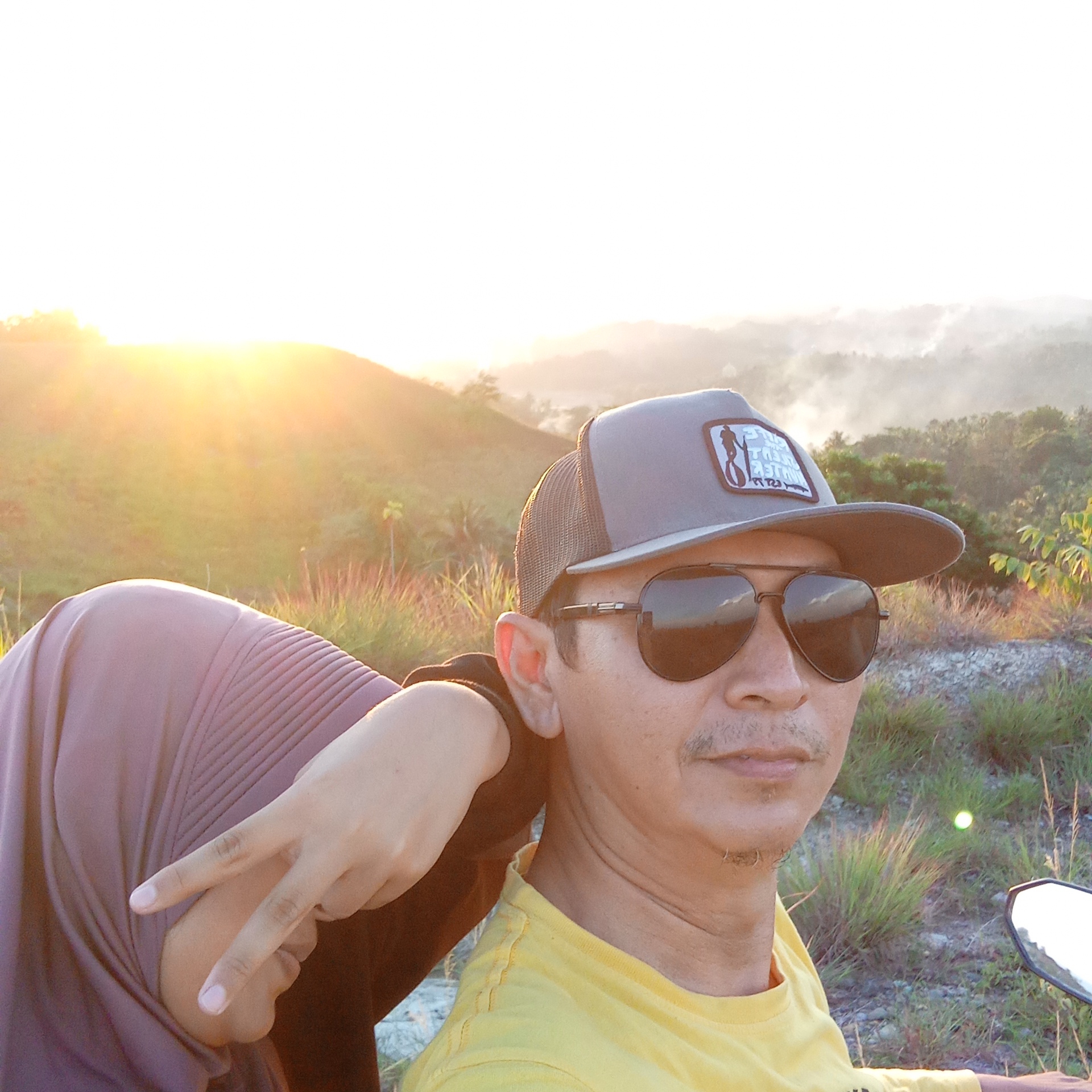 In Lhokseumawe, Indonesia, a man wearing a grey trucker hat and aviator sunglasses is photographed in a close-up selfie. He is wearing a yellow t-shirt. To his left, a woman wearing a purple hijab rests her head on his shoulder and makes a peace sign with her hand. The background depicts a landscape of rolling hills and trees bathed in the warm glow of a sunset. The scene suggests a casual outing, possibly a motorcycle ride, given the visible motorcycle mirror in the lower right corner.
