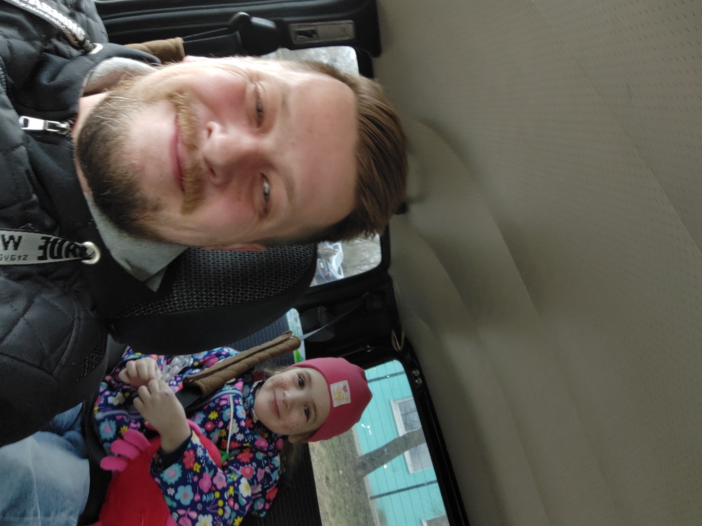 A man and a young girl are in a car. The man is in the driver's seat, and the girl is in the back seat. The man is smiling and looking at the camera. The girl is smiling and looking at the camera. The location is in the city of Chaikovsky, Russia. The car is parked on a street. There are trees and buildings in the background.