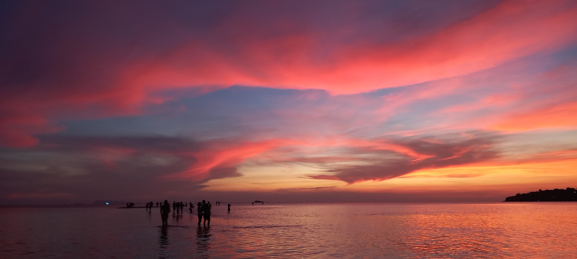 The image shows a sunset scene over the ocean in Baan Sri Thanu, Thailand. The sky is filled with vibrant colors of red, orange, pink, and purple, with clouds scattered throughout. The water reflects the colors of the sky. There are several people wading in the shallow water near the shore. A small, dark landmass is visible on the right side of the image.
