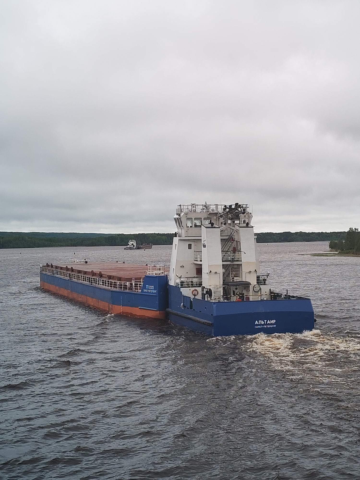 A blue and orange cargo ship, identified as "АЛЬТАИР" (Altair) from Saint Petersburg, Russia, travels on a wide body of water under a cloudy sky. The vessel consists of a flat barge section and a multi-level control tower structure. The water surface is moderately rough with visible wake from the ship's movement. In the background, wooded shorelines can be seen on both sides of the water, with another distant vessel visible in the center. The photograph does not confirm it being located in Анхимово, Russia, but this information is given in the prompt.