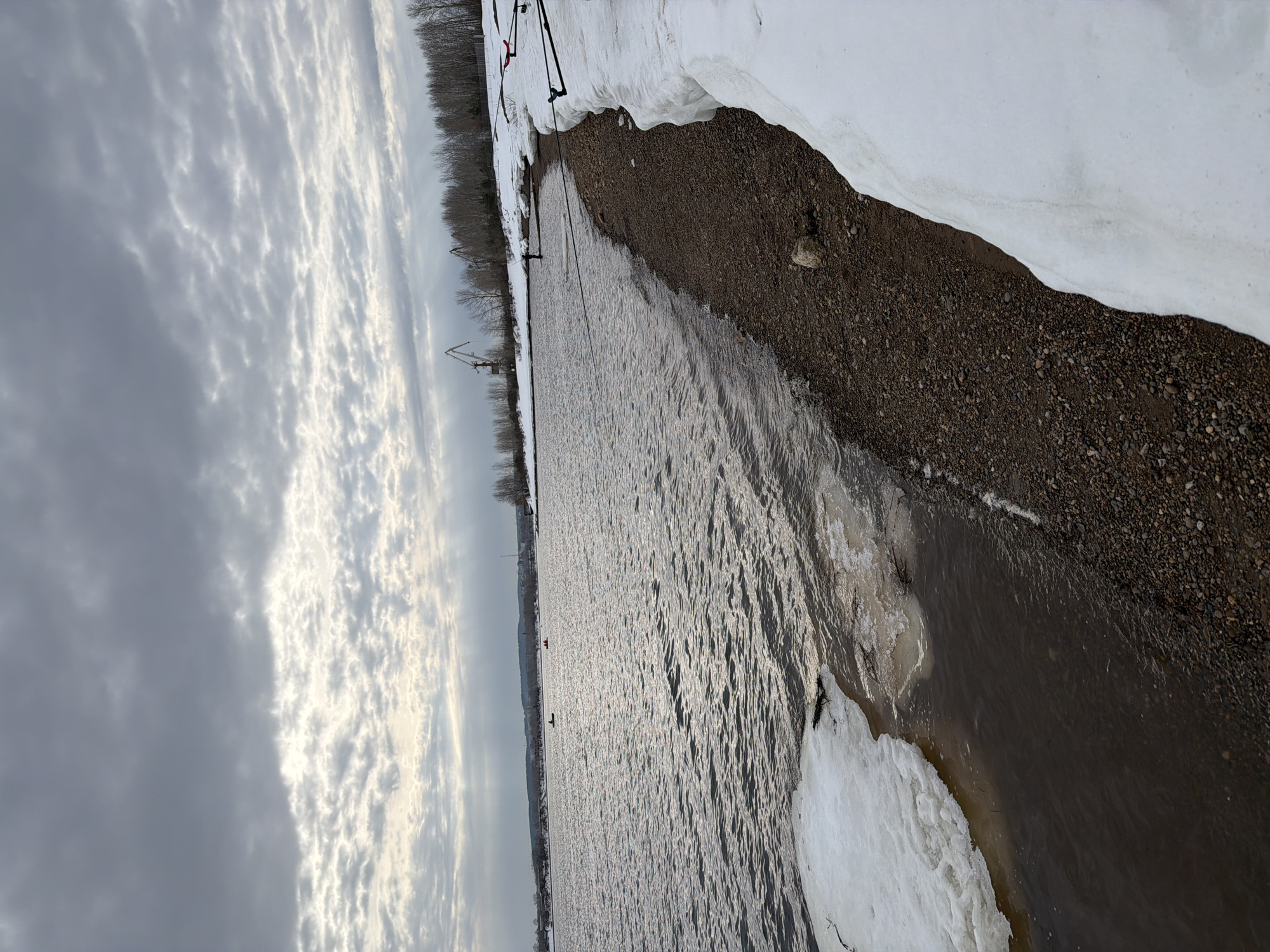 The image depicts a landscape in what appears to be the city of Chaikovsky, Russia, during late winter or early spring. The scene is dominated by a body of water, likely the Kama River, with shimmering, textured surface due to sunlight reflecting off the ripples. Along the riverbank, there is a strip of land covered with dark gravel or pebbles, and patches of melting snow and ice.
On the far side of the river, a line of bare trees stands against the horizon under a sky filled with striated clouds. There are some poles that could be for kitesurfing.