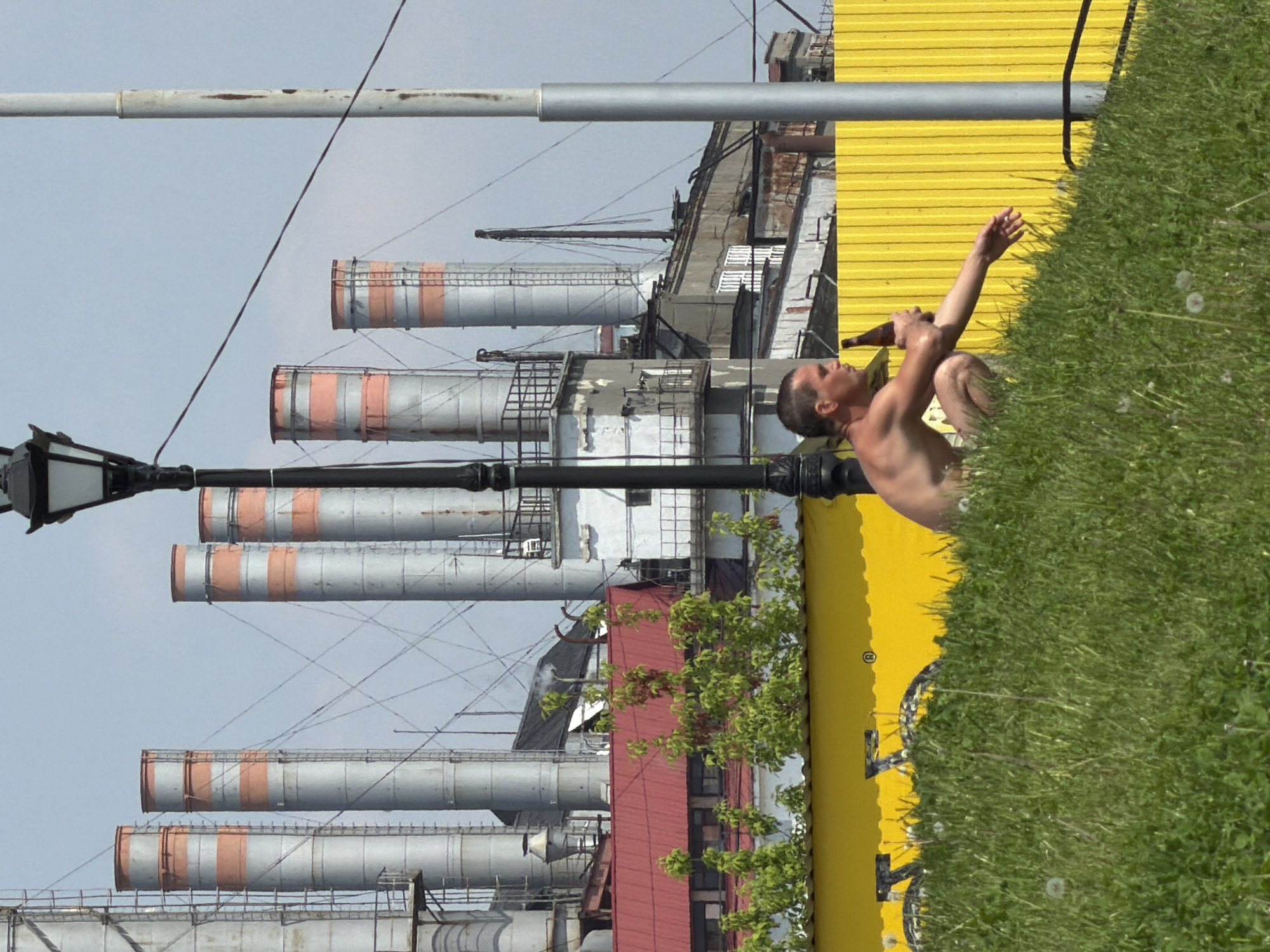 A shirtless man crouches in grass beside a yellow wall, holding a bottle to his mouth, with his right arm raised. In the background are several industrial smokestacks with orange stripes, along with buildings and a lamppost. This is located in Saratov, Russia.