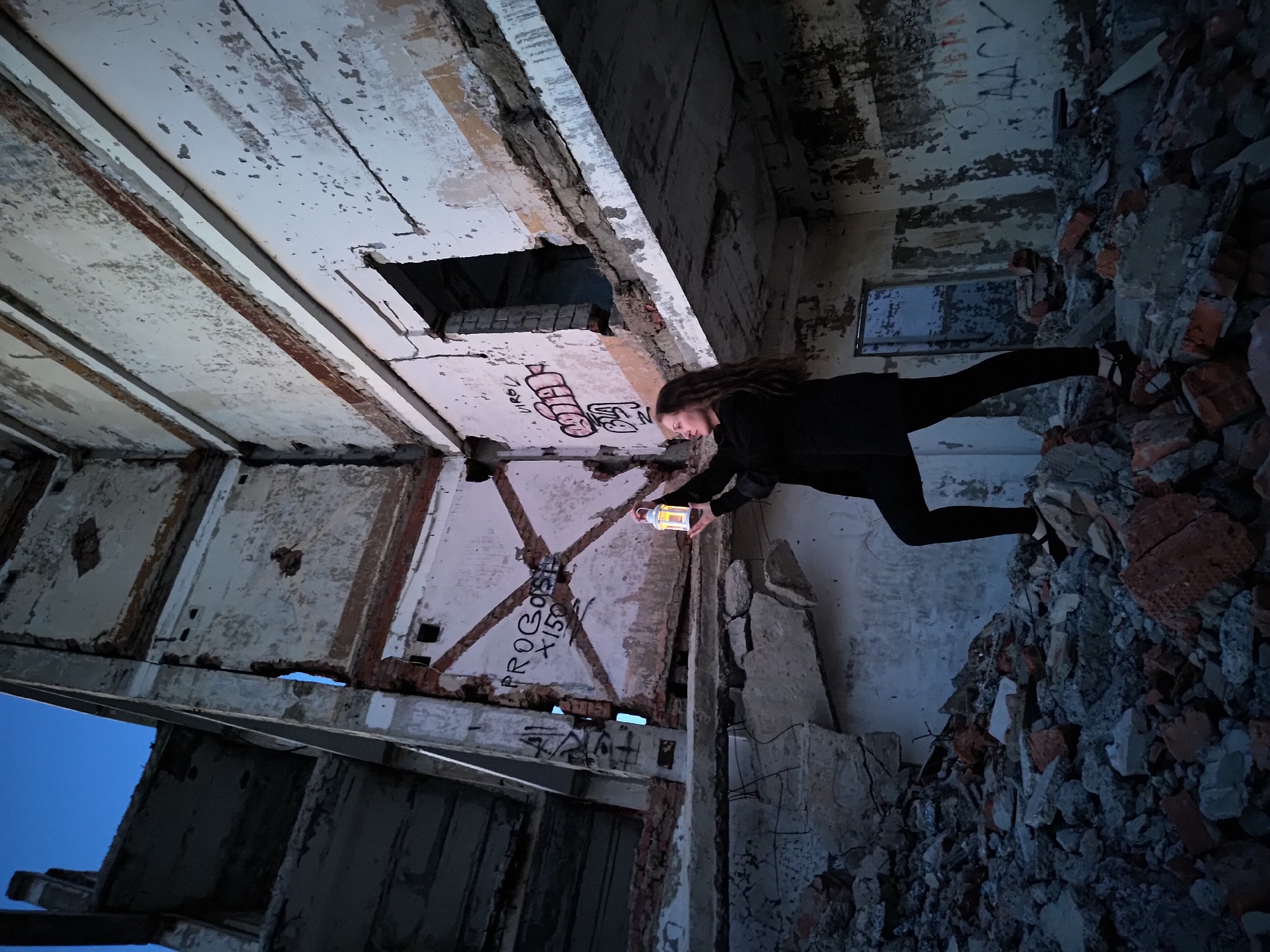 A woman dressed in black is holding a lantern inside the decaying interior of a brick and concrete building. She is positioned on the right of the frame amid a pile of rubble, and the building's remains surround her. The architecture exhibits severe disrepair, with broken walls, exposed beams, and peeling paint. Graffiti is visible on several surfaces. The setting sun gives off a blue glow in the areas that are not lit by the lantern. It is not possible to confirm the location in the city of Марковский, Russia.