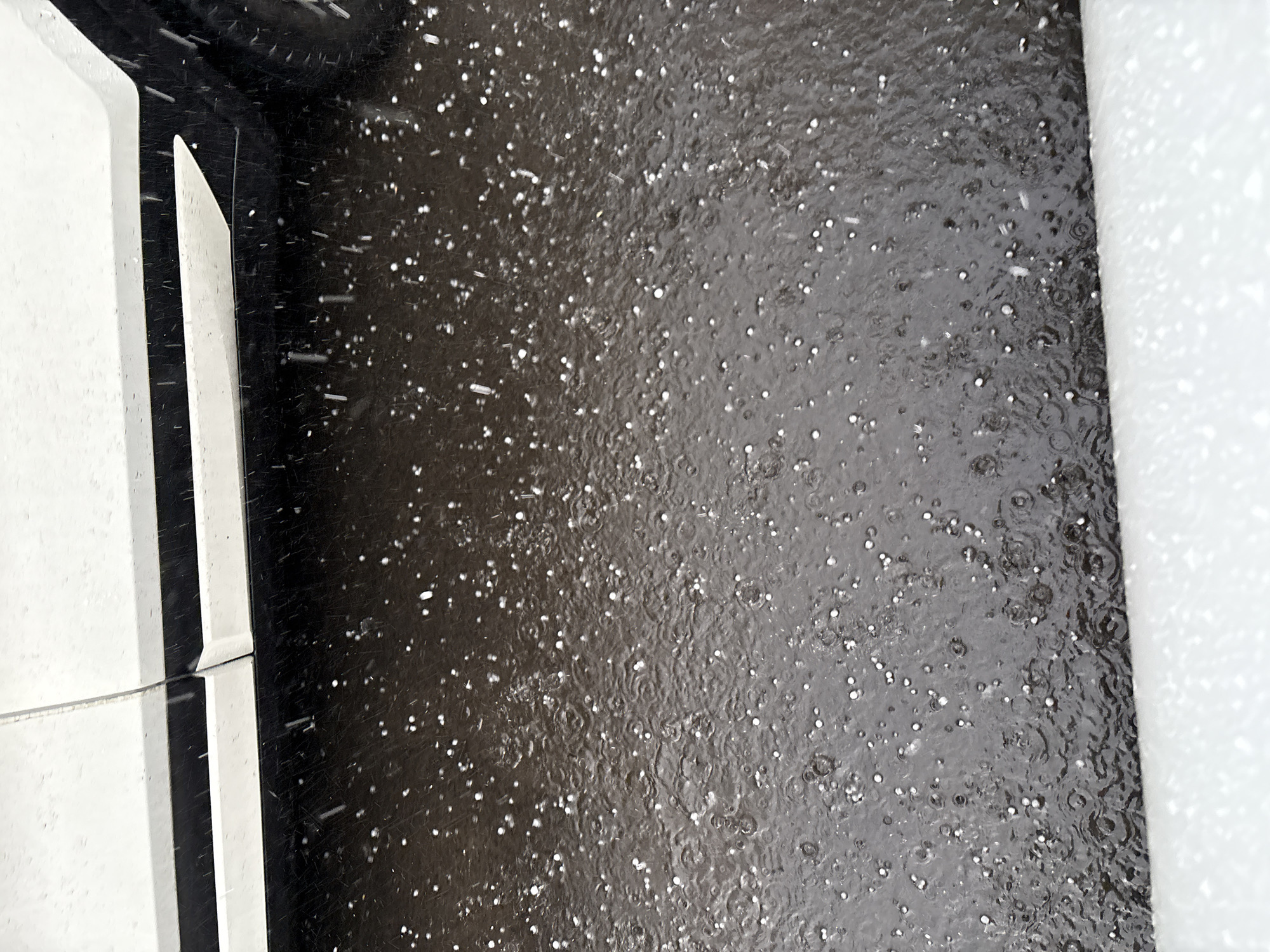 The image depicts a close-up, high-angle view of hail falling on a dark, wet asphalt surface next to the lower body of a white vehicle. The hail appears as small, white, spherical particles scattered across the ground. The asphalt is reflective, showing ripples and splashes caused by the impact of the hail and raindrops. The white vehicle, visible in the upper portion of the frame, has a dark accent line running along its lower edge. The foreground is occupied by a blurry white surface, possibly a curb or snowdrift. The lighting suggests an overcast or dimly lit day. The scene is devoid of any visible people or animals. The presence of hail indicates a specific weather event occurring at the time the photograph was taken. Without additional information, the precise location within Ust-Ilimsk, Russia, cannot be determined.