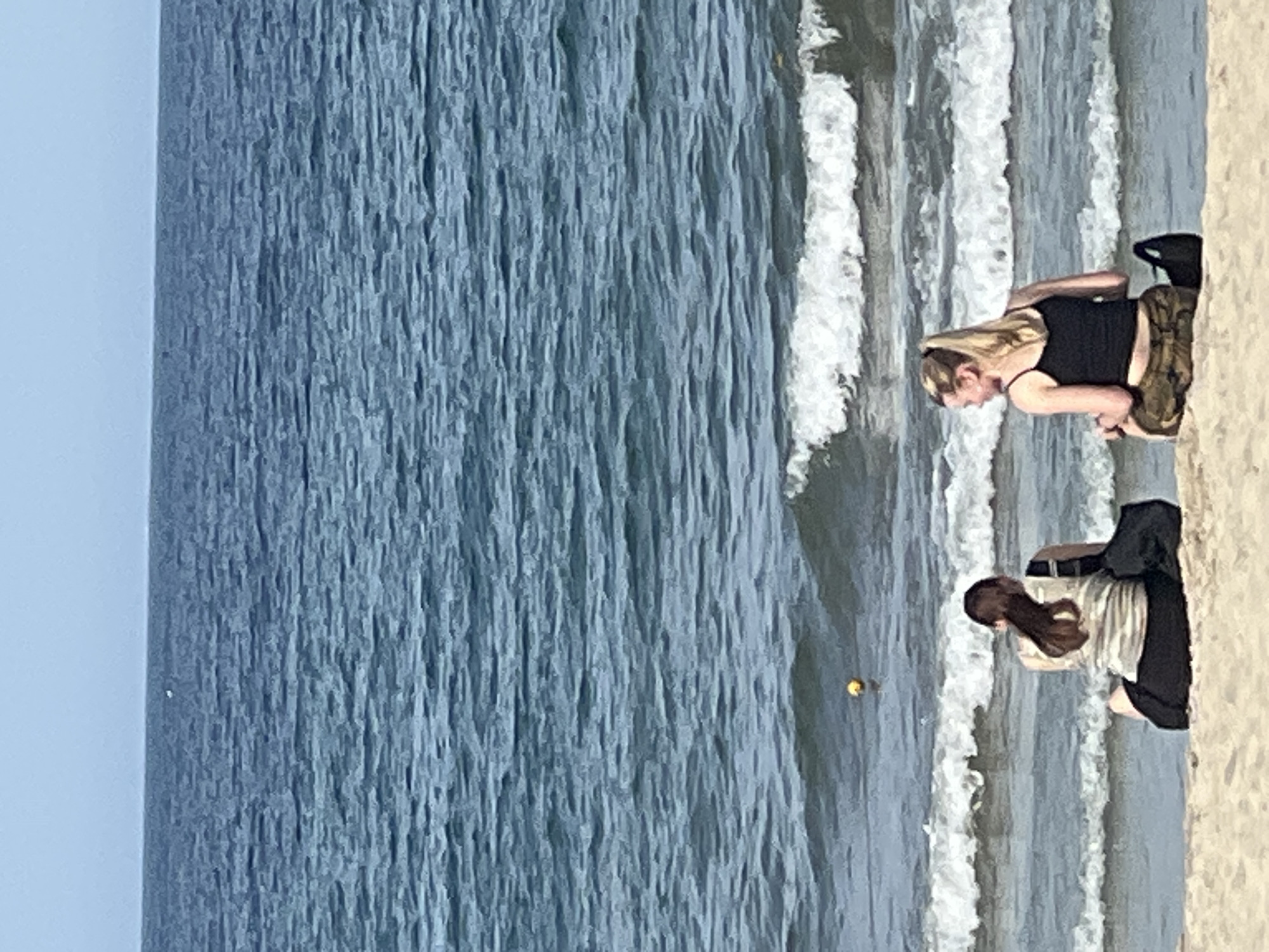 The image depicts a serene beach scene on a sunny day. Two young women are seated on the sand, facing the ocean. The woman on the left has long, dark hair and is wearing a striped tank top and dark shorts. She has a black bag beside her. The woman on the right has blonde hair tied in a ponytail and is wearing a black tank top and camouflage shorts. She also has a black bag next to her. The ocean is a deep blue with white waves gently rolling towards the shore. In the distance, a small yellow buoy is visible in the water. The sky is a pale blue, indicating clear weather. The location is likely My Khe Beach in Da Nang, Vietnam, known for its long stretches of sand and clear waters.