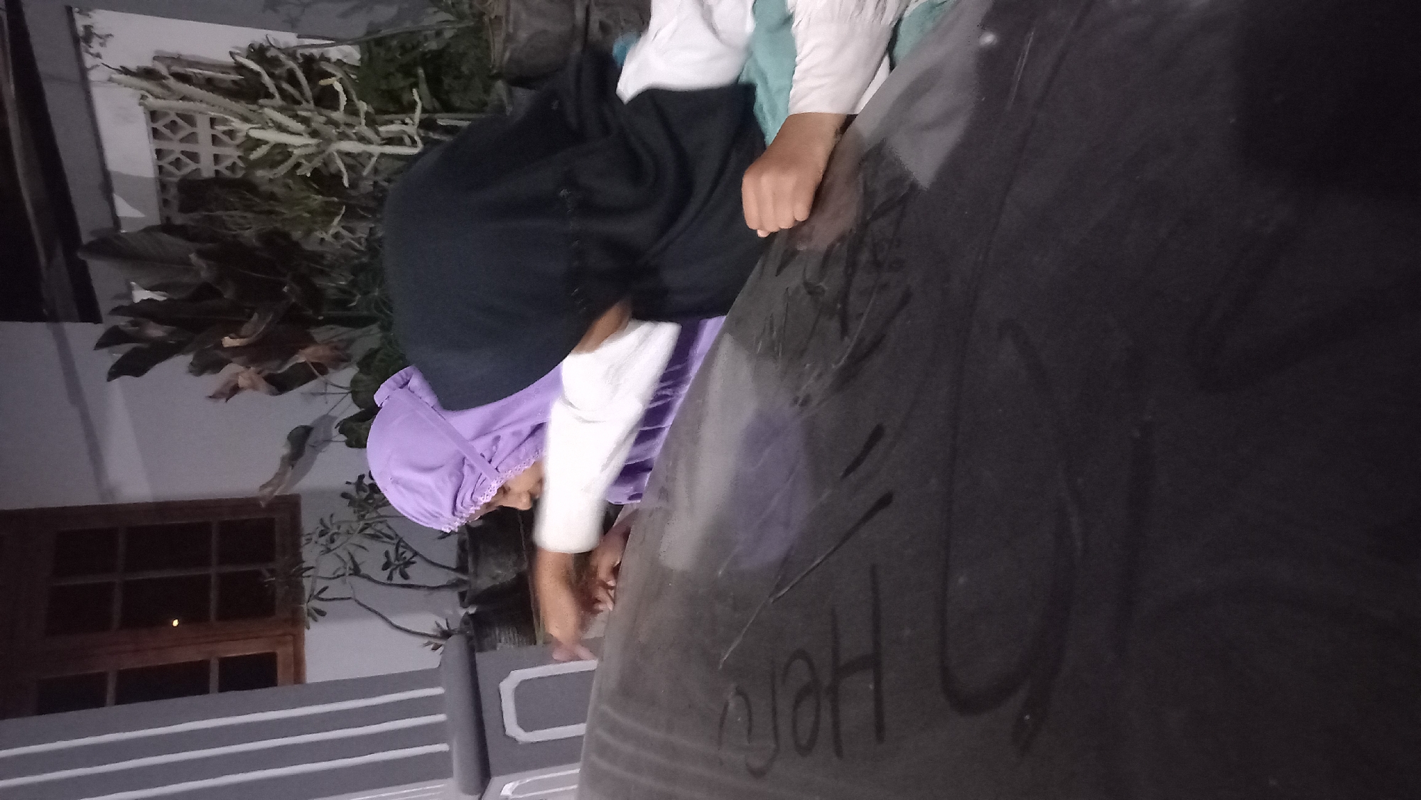 The image captures a nighttime scene in Pare, Indonesia, focusing on two individuals, likely children or young teenagers, interacting with a dark, smooth surface. The environment suggests an outdoor or semi-outdoor setting, possibly a patio or a covered area, with a building structure visible in the background.
One individual, wearing a purple headscarf, is positioned on the left side of the frame, with their right hand reaching out towards the surface. Their body is angled downwards, indicating they are leaning over. The other individual, wearing a black headscarf, is partially visible on the right, also leaning forward, with their left hand resting on the surface.
The dark surface in the foreground displays visible markings or writing. On the left side of this surface, the word "HERO" is clearly written, seemingly with a finger or a similar object, creating a contrast with the dark background. There are also other faint, indistinct markings on the surface.
In the background, a window with wooden frames is visible on the left, illuminated from within. Adjacent to the window are potted plants, one with thin branches and leaves, and another with darker, broader leaves. Further to the right, a decorative grill pattern is visible on a wall, and more plants are present, some appearing spiky and pale, others darker and leafy. The overall lighting suggests it is dark, with ambient light sources illuminating certain areas. The scene implies a moment of casual interaction and play in a residential or public space in Pare.