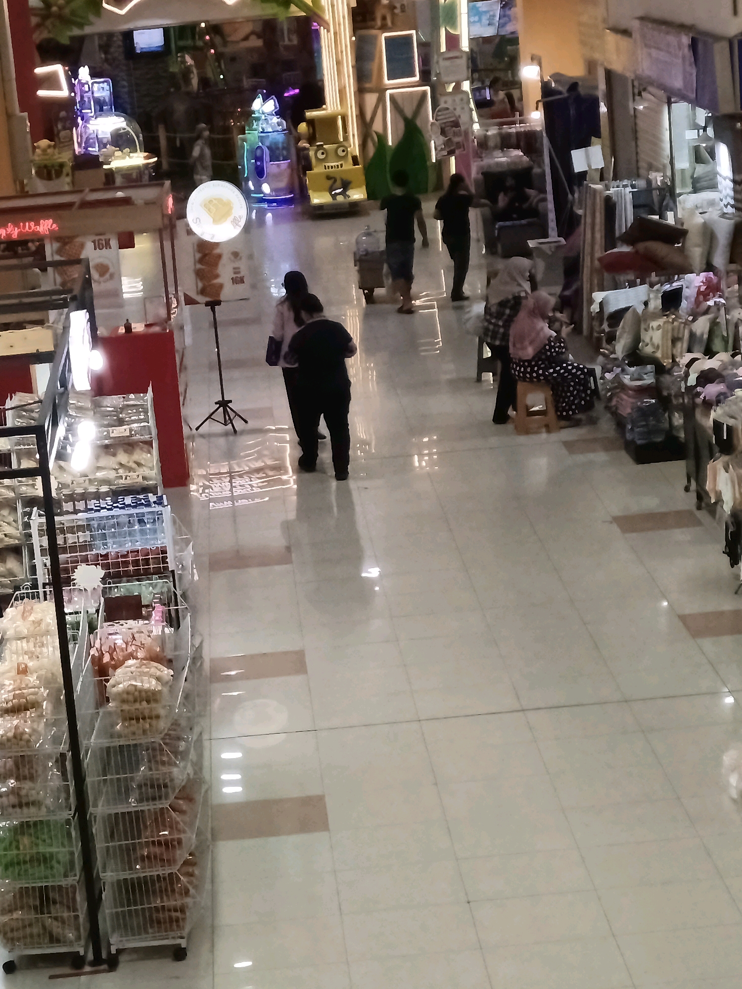 The image captures a bustling indoor market or mall in Central Jakarta, Indonesia, from a high-angle perspective. The brightly lit environment features tiled flooring that reflects the overhead lights. In the foreground, a rack filled with various bagged snacks or food items is prominently displayed. To the left, a red stall with signage indicates a food vendor, possibly selling waffles, with pricing of "16K" visible.
Several individuals are present in the scene. Two people walk away from the camera towards the left, while further back, a group of three individuals are seen, two of whom are walking and one is partially obscured. A woman wearing a headscarf is seated on a stool on the right, near a display of textiles and clothing items.
The background reveals more of the market's offerings, with various stalls and displays of goods. Entertainment or game machines, illuminated with colorful lights, are visible in the upper left section of the image. The overall impression is one of a lively commercial space with a variety of products and shoppers.