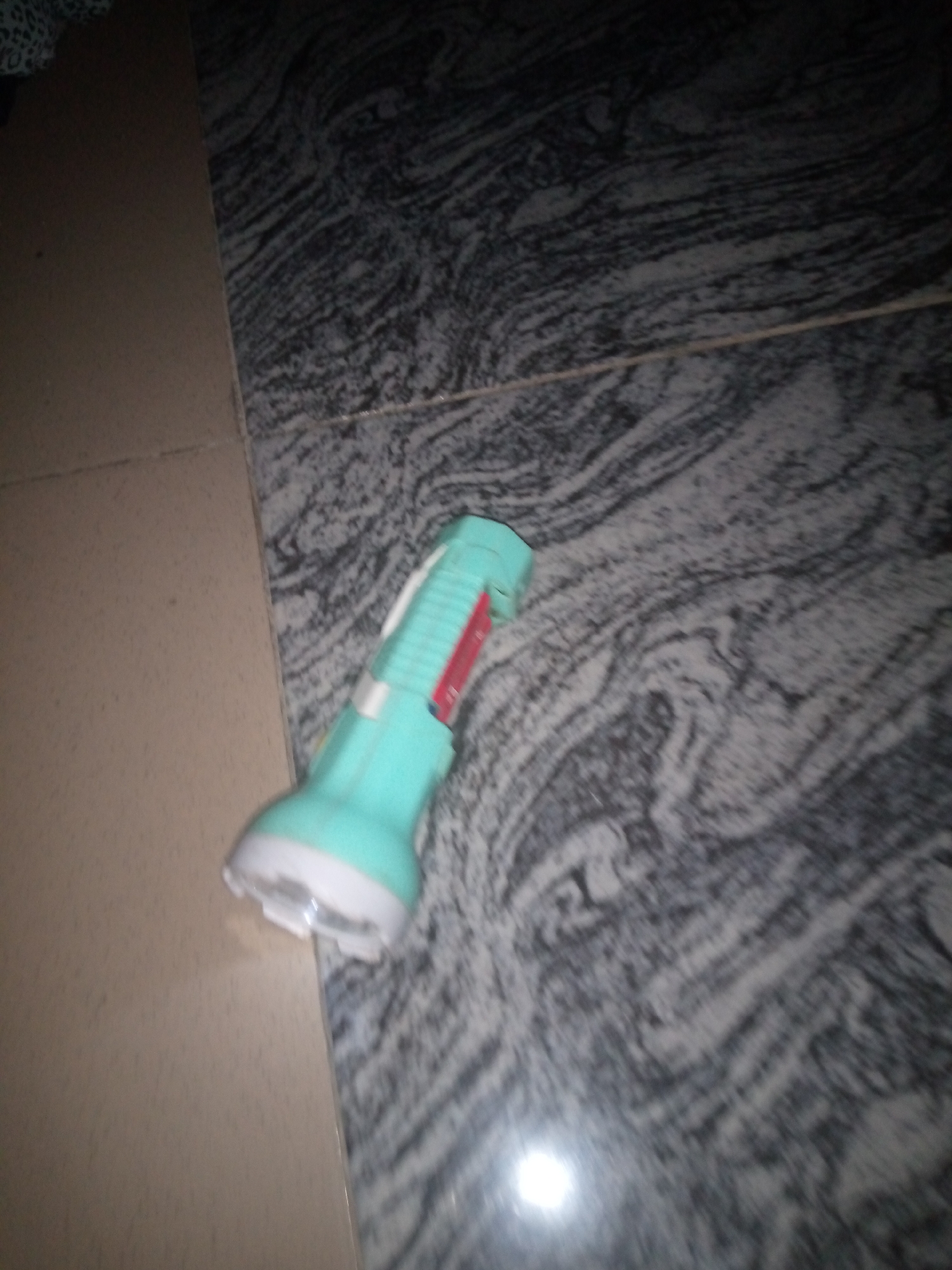 A turquoise and white flashlight with a red accent is lying on a dark, patterned surface, possibly marble, which appears to be a floor. To the left of the flashlight, a beige tiled surface is visible, meeting the dark surface at a right angle. The scene is dimly lit, with a bright reflection visible on the dark surface. There are no people or animals present in the image, and no discernible action is taking place. The flashlight appears to be an ordinary household object. Given the context provided, this image was taken in the city of Jalingo, Nigeria.